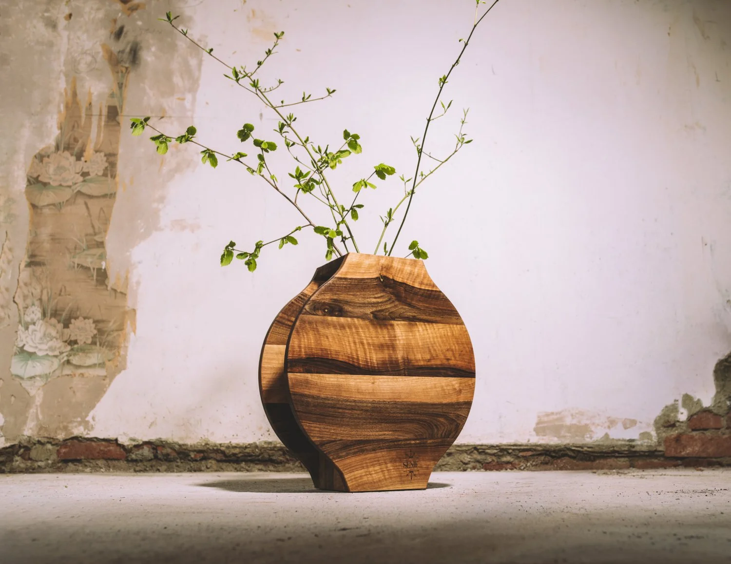 A wooden vase with green leafy branches against a weathered wall.
