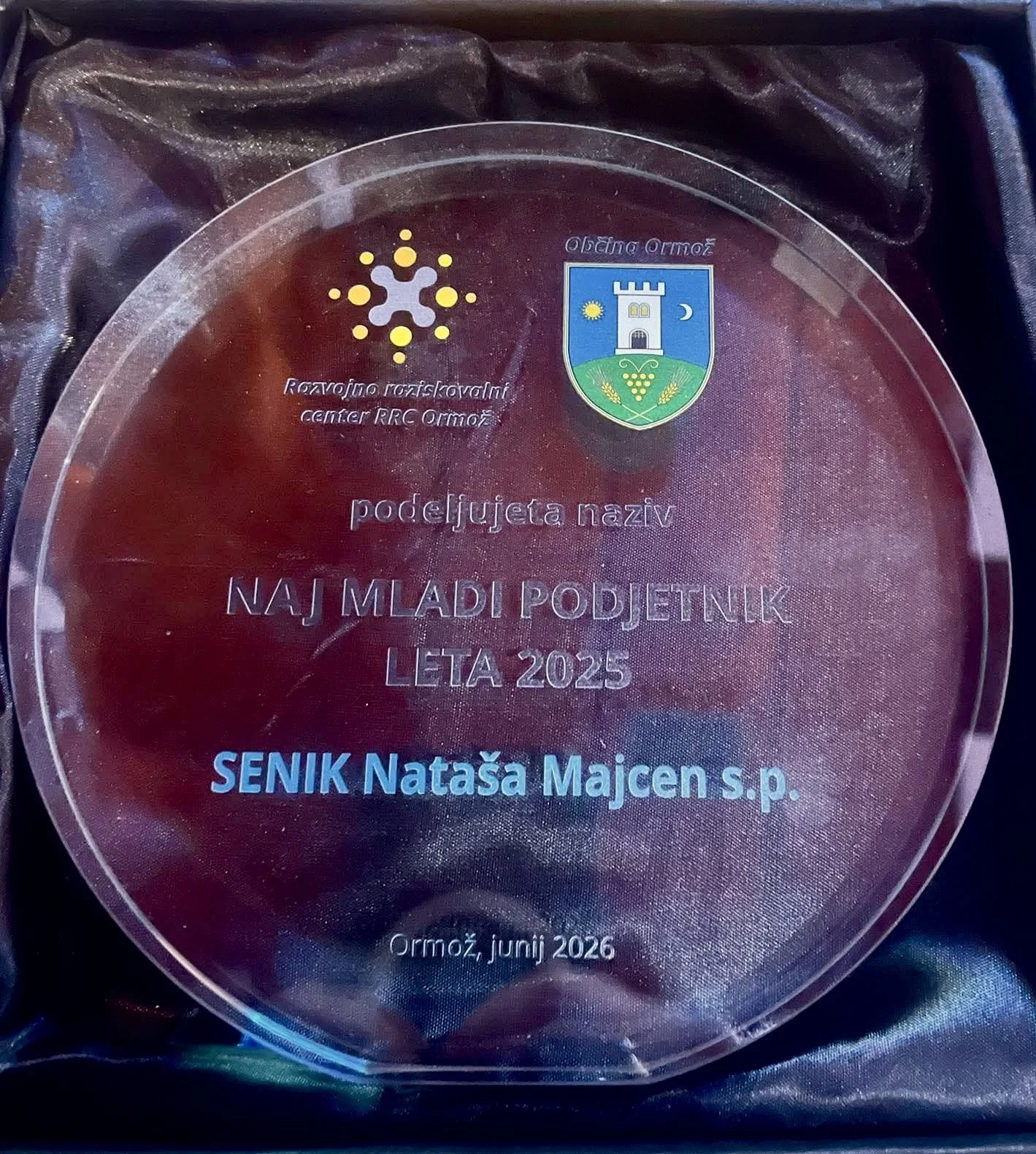 A circular glass award with inscriptions in Croatian, featuring a crest with a tower, sun, moon, and wheat. The award is for the 'Naj Mladji Podjetnik Leta 2025,' awarded to SENIK Nataša Majcen in June 2026 in Ormož.