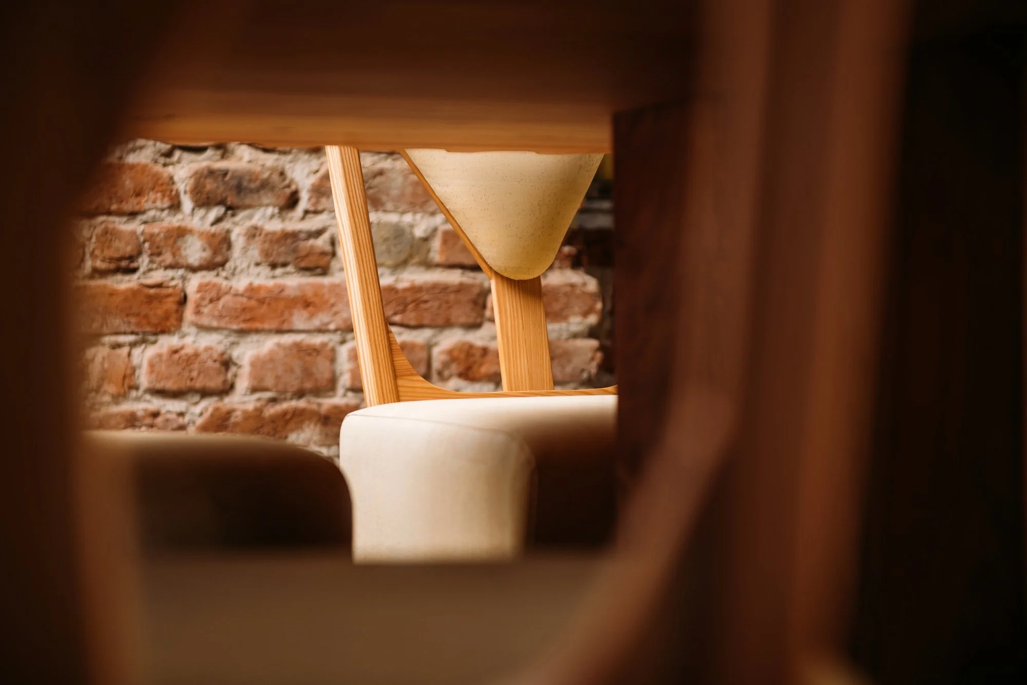 View of a modern wooden chair with a beige cushion, seen through a small opening between wooden panels, with a red brick wall in the background.