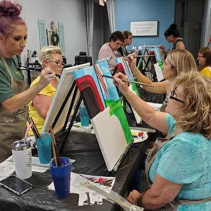 People participating in a group painting activity, sitting at a long table and painting on canvases with different colors. Some are wearing glasses and aprons, with painting supplies around them.