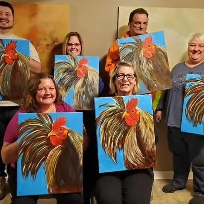Six people posing with completed rooster paintings they created, each holding their own artwork painted on canvases, smiling indoors.
