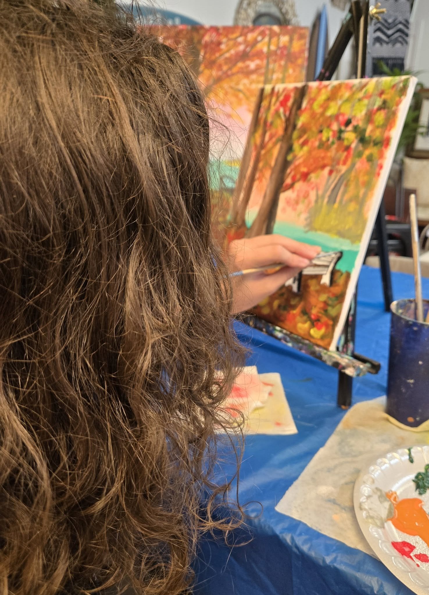 A person with long, curly hair painting fall scenery on a canvas, featuring trees with orange and yellow leaves and a blue stream, at an art class.