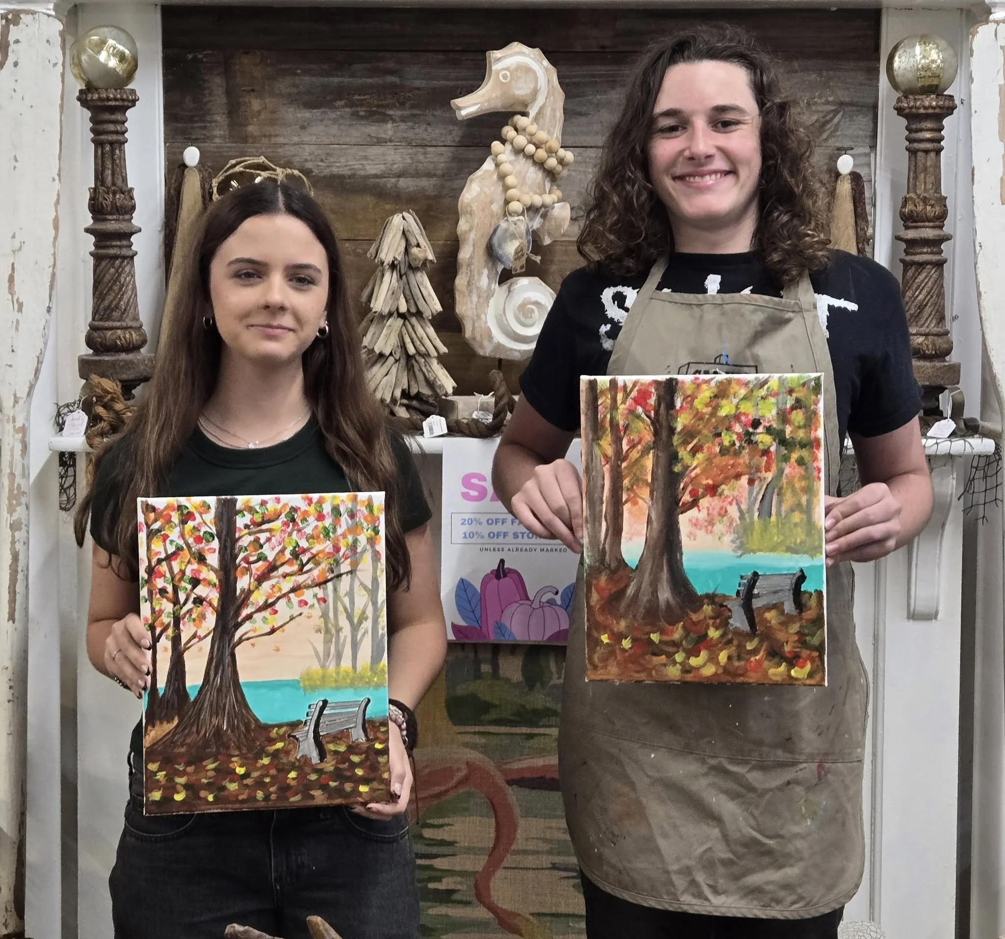 Two young women holding paintings of autumn trees with colorful leaves and a bench by a lake, standing in front of a wooden background with art and decor.