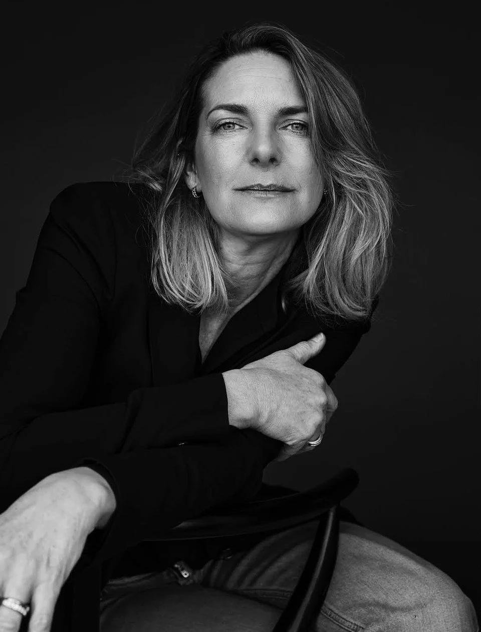 Zwarte en witte portretfoto van een vrouw met lange haar, die rustig kijkt en een zwarte jas draagt, zittend op een stoel voor een zwarte achtergrond.