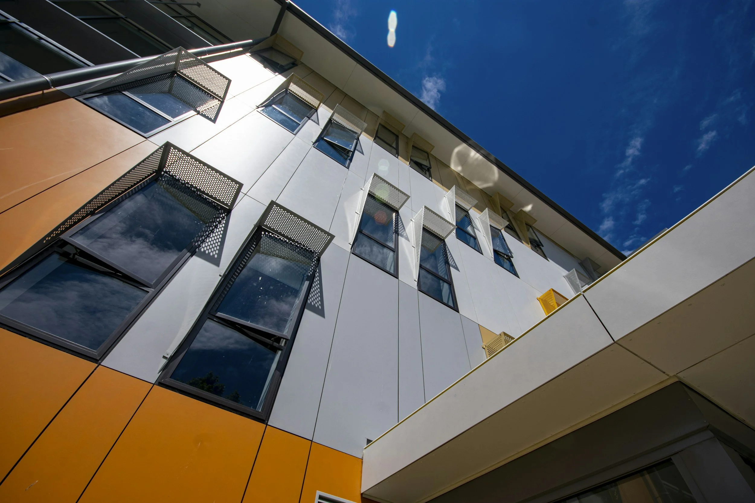Low-angle view of a modern building with windows featuring small sunshades, against a bright blue sky with a few clouds.