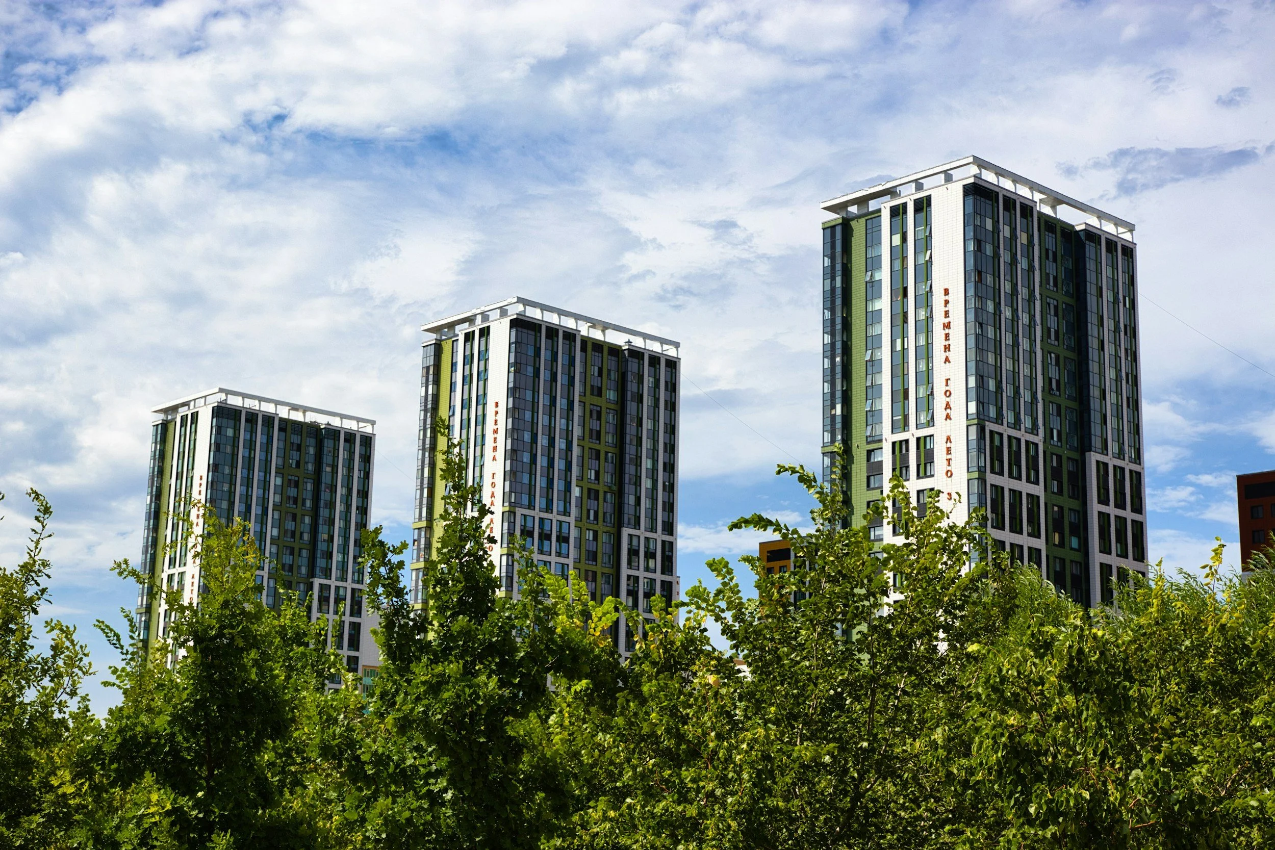Three modern high-rise buildings with green and white exteriors under a partly cloudy sky, with trees in the foreground.