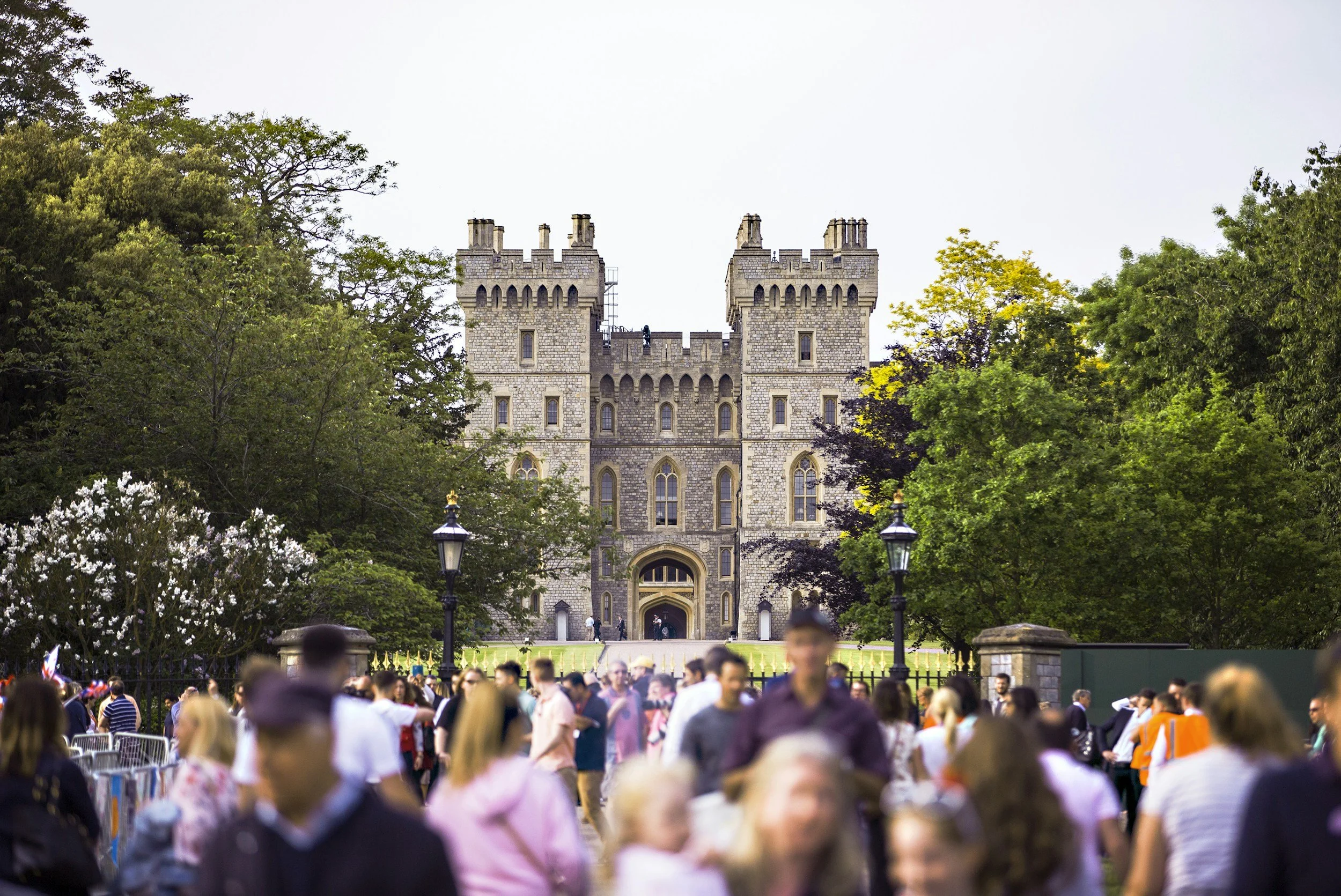 The image shows a large crowd gathered in front of a castle-like building surrounded by trees. The building has towers and arched windows, resembling a historical castle or estate.