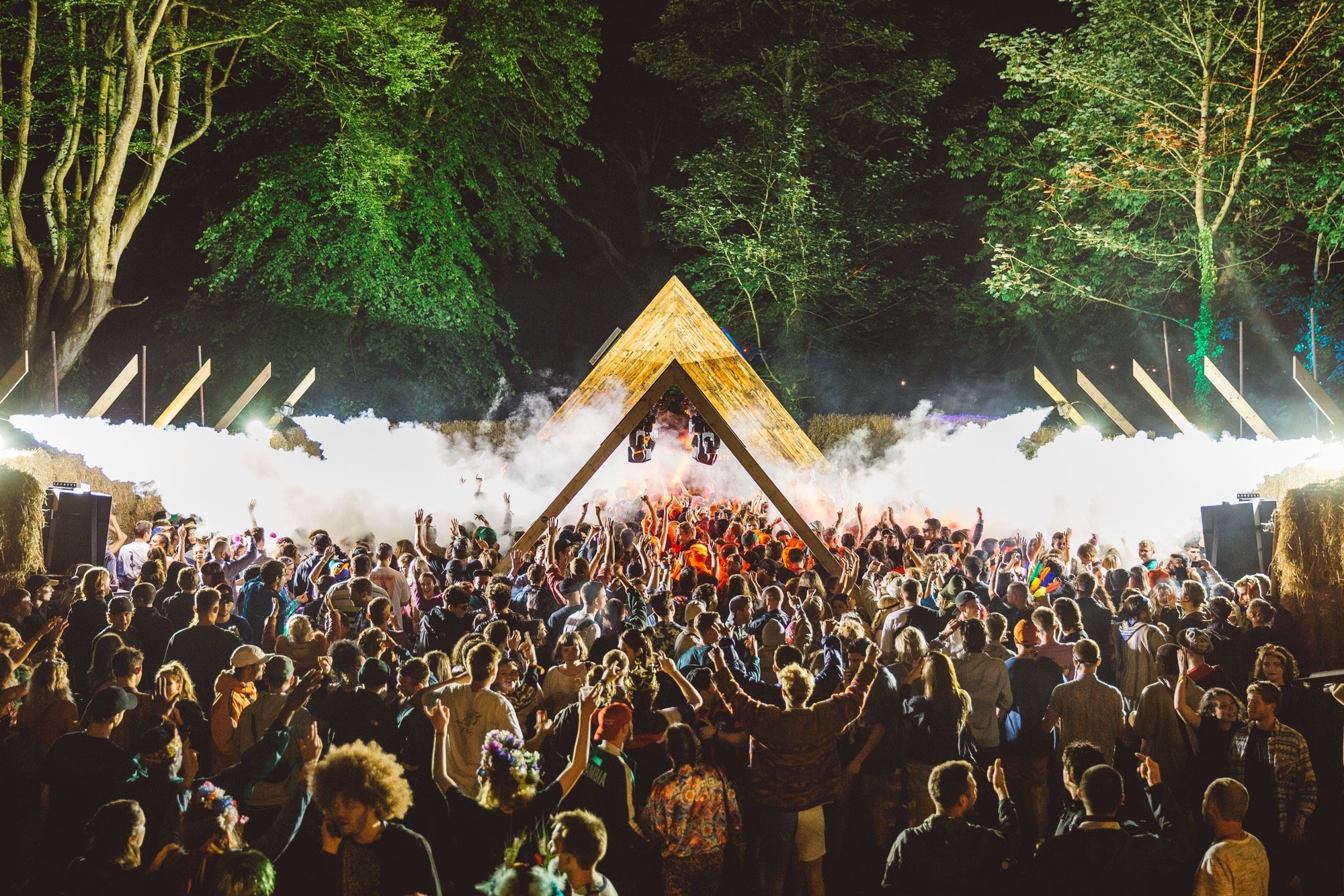 A large crowd of people at an outdoor night concert or festival, with a stage featuring a wooden triangular structure, surrounded by trees and illuminated with bright lights and fog effects.