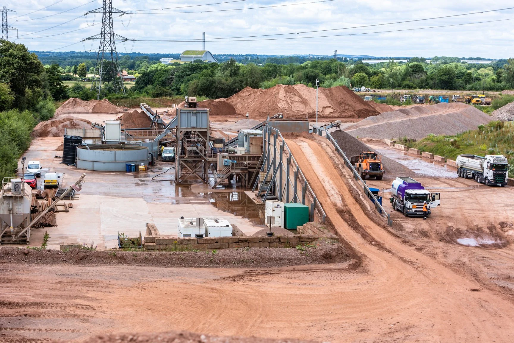 Construction site with dirt mounds, construction vehicles, and industrial equipment, surrounded by greenery and power lines in the background.
