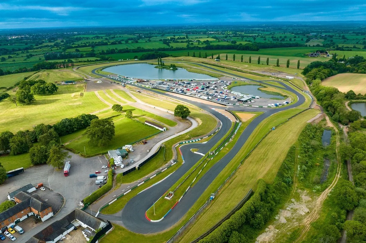 Aerial view of a race track with a winding asphalt course, surrounded by green fields, water bodies, and a parking lot filled with numerous vehicles.
