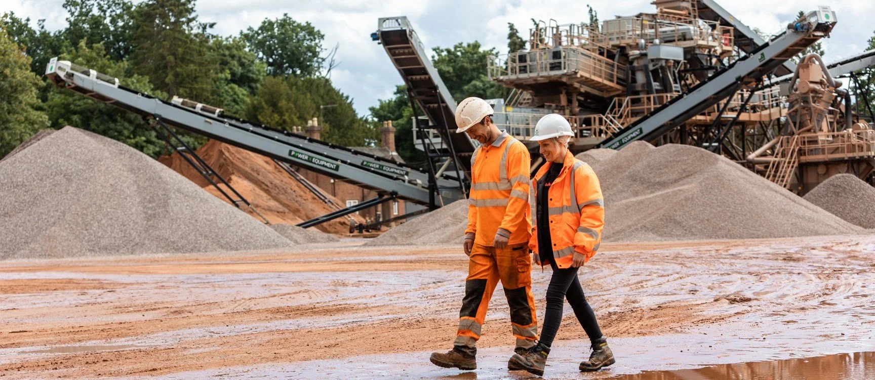 Two construction workers, a man and a woman, wearing orange safety jackets and white hard hats, walking on a muddy construction site with piles of gravel and large industrial machinery in the background.