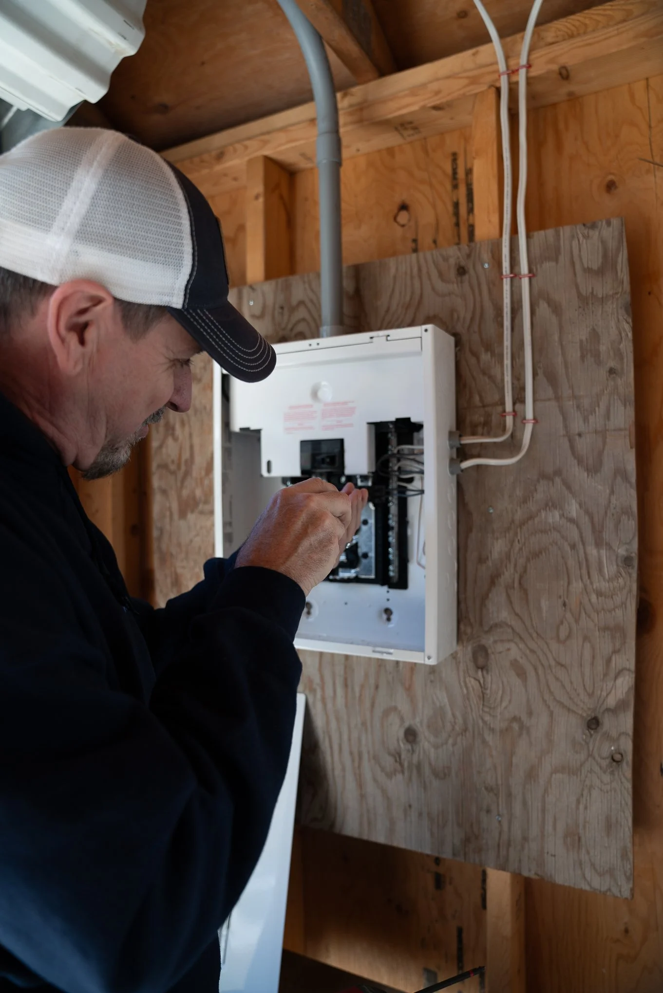 A man wearing a baseball cap and black jacket working on electrical wiring inside an electrical box on a unfinished wooden wall.