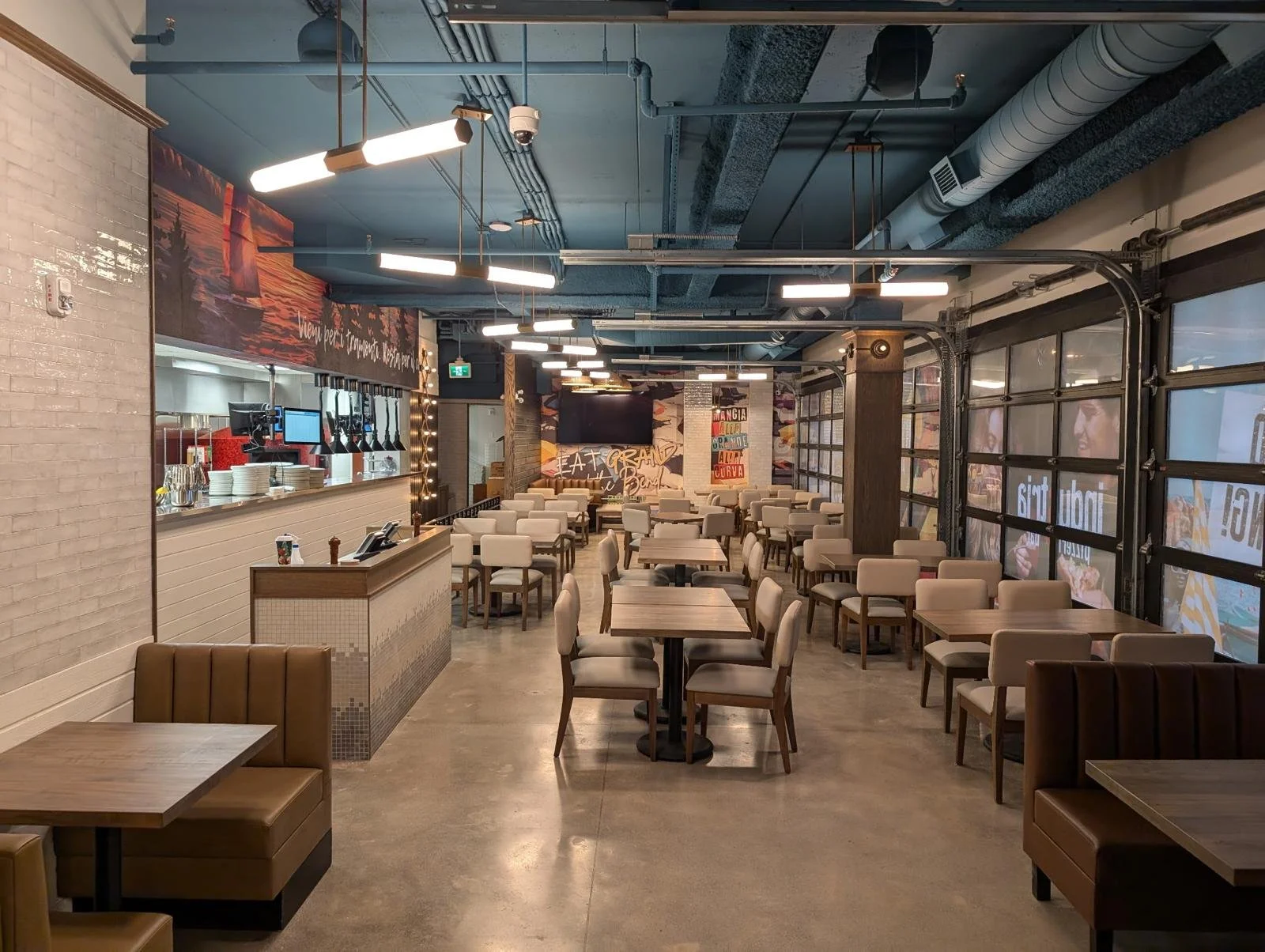 Interior of a modern restaurant or cafe with wooden tables and beige upholstered chairs, a counter on the left with plates and coffee machines, exposed brick and painted walls, large windows on the right with a garage-style door, and decorative lighting on the ceiling.