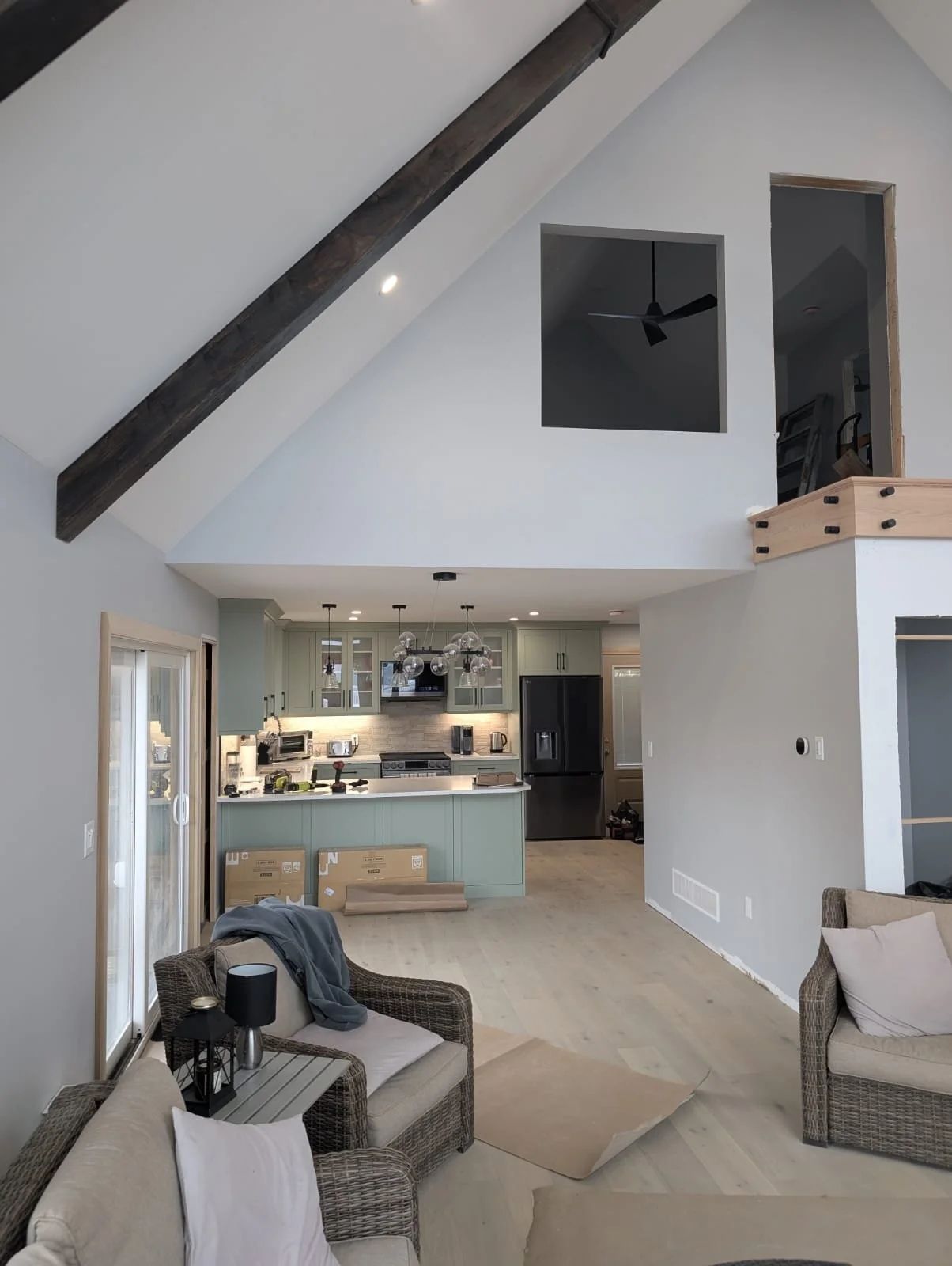 Living room and kitchen with high vaulted ceiling, exposed dark wood beam, and large windows, featuring light-colored walls and modern furniture.