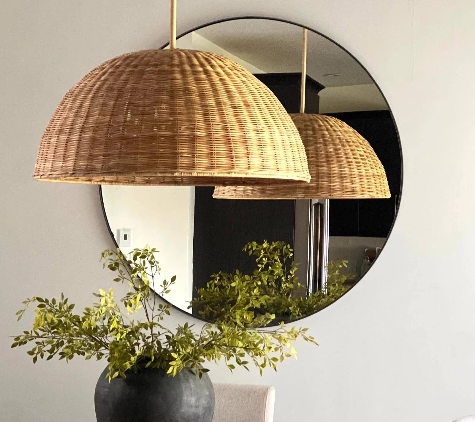 Wicker globeshaped hanging lamps, four hanging from the ceiling, with a round mirror on the wall behind them. In the foreground, a vase with green leafy plant on a dining table.