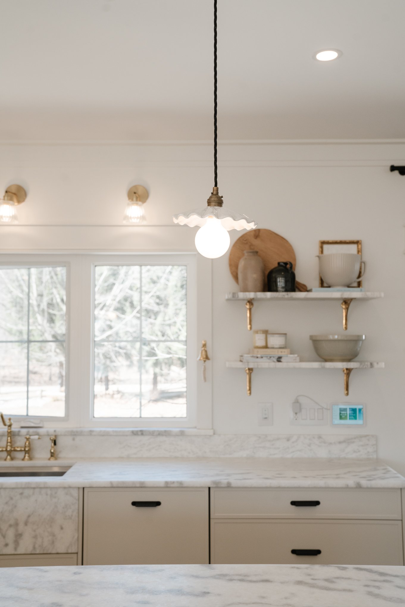 Modern kitchen with a marble countertop, white cabinets, a window with trees outside, and open shelving with vases and bowls. A pendant light hangs from the ceiling.