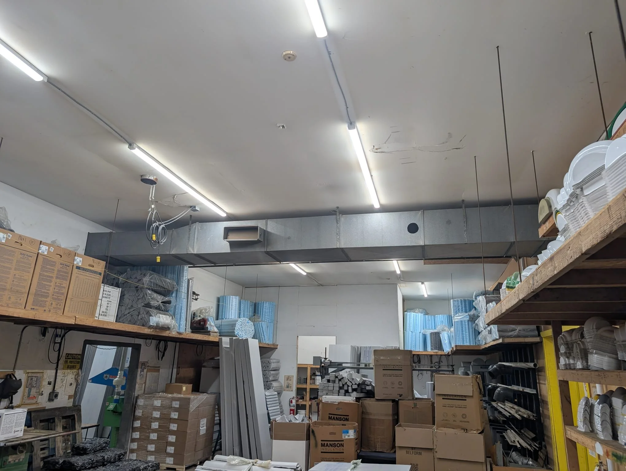 View of a hardware store aisle with shelves stocked with boxes, rolls of insulation, and various building supplies. The ceiling has fluorescent lighting and some visible wires and fixtures.