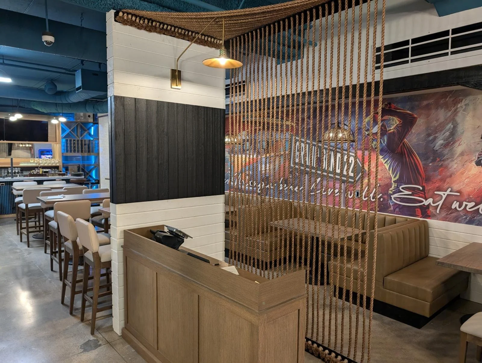 Interior of a modern restaurant with beige chairs and booths, wooden accents, decorative rope divider, and a colorful mural wall depicting a woman and dynamic paint splashes.