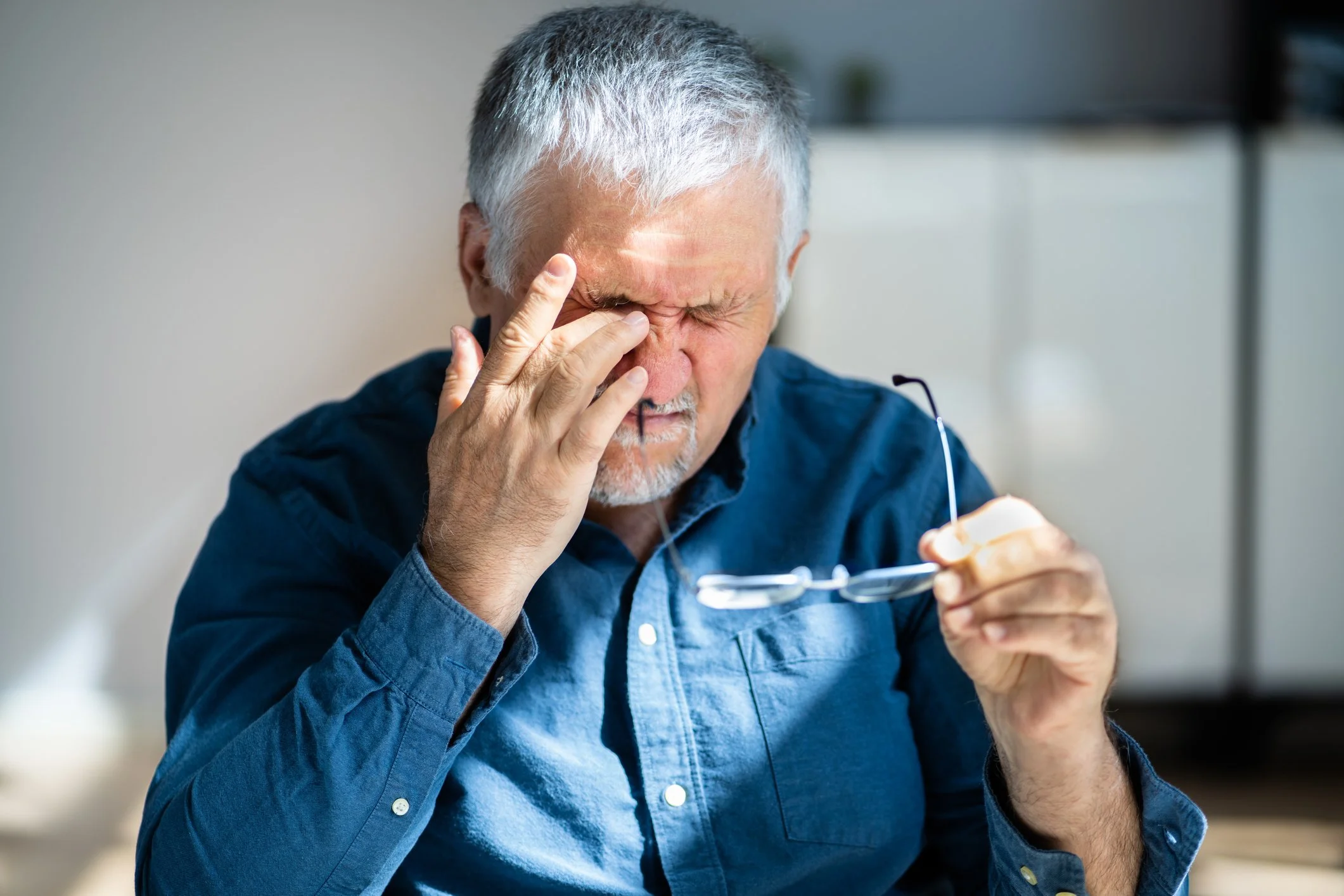 An elderly man with gray hair and a beard, wearing a blue shirt, holds eyeglasses in one hand while rubbing his forehead with his other hand, showing signs of discomfort or headache.