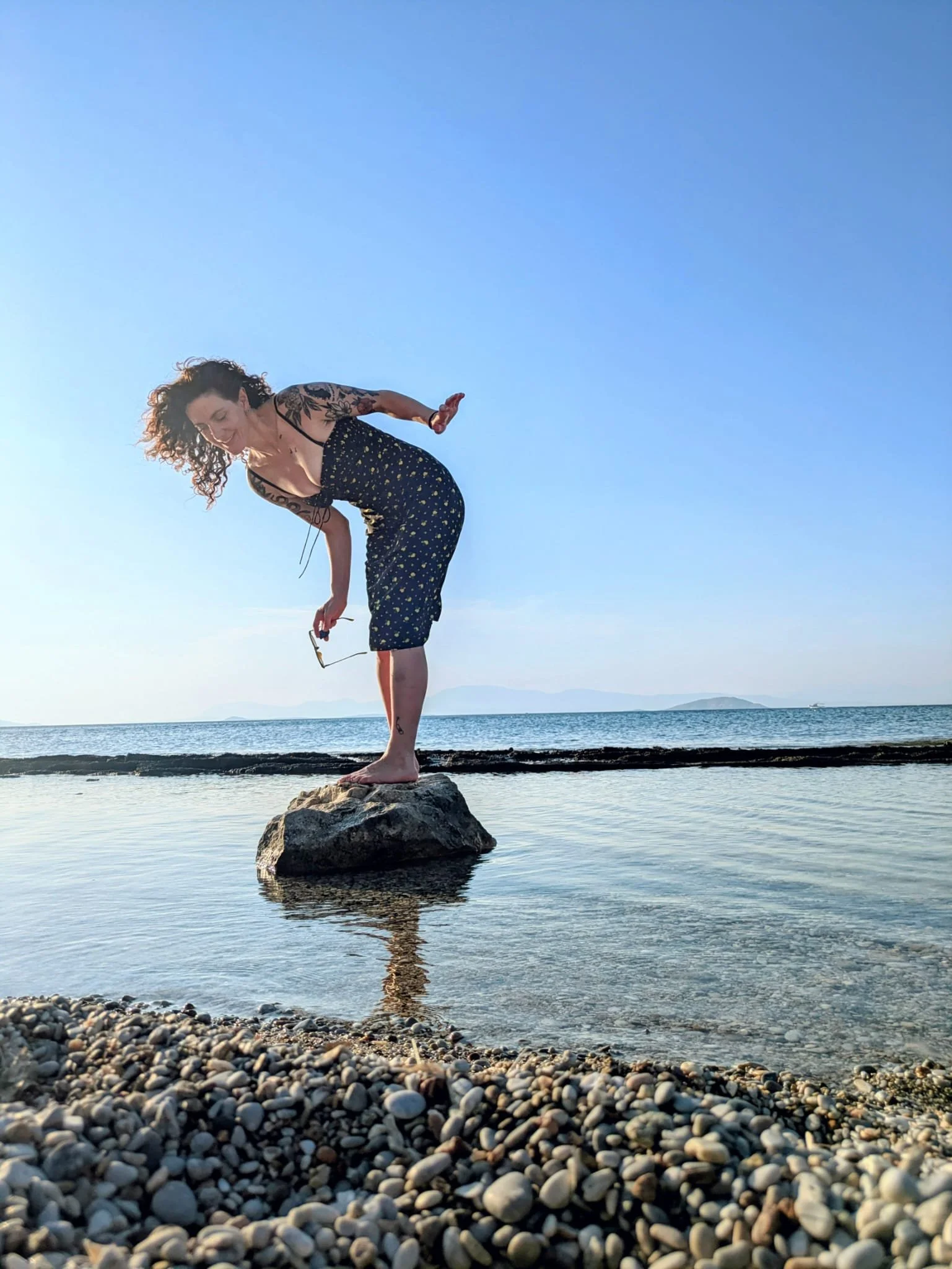 Celine, all natural, beautiful companion, escort, sex worker in London. She wears a classy black silk dress and her long sexy hair curls naturally to her shoulders. She is balancing on a rock in the ocean, holding a pair of designer sunglasses.