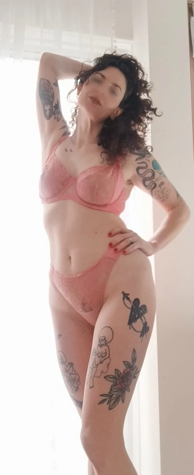 A woman with curly black hair and tattoos on her arms and legs is posing in pink lace lingerie with one hand on her hip and the other behind her head, standing in front of a light-colored wall with window in the background.