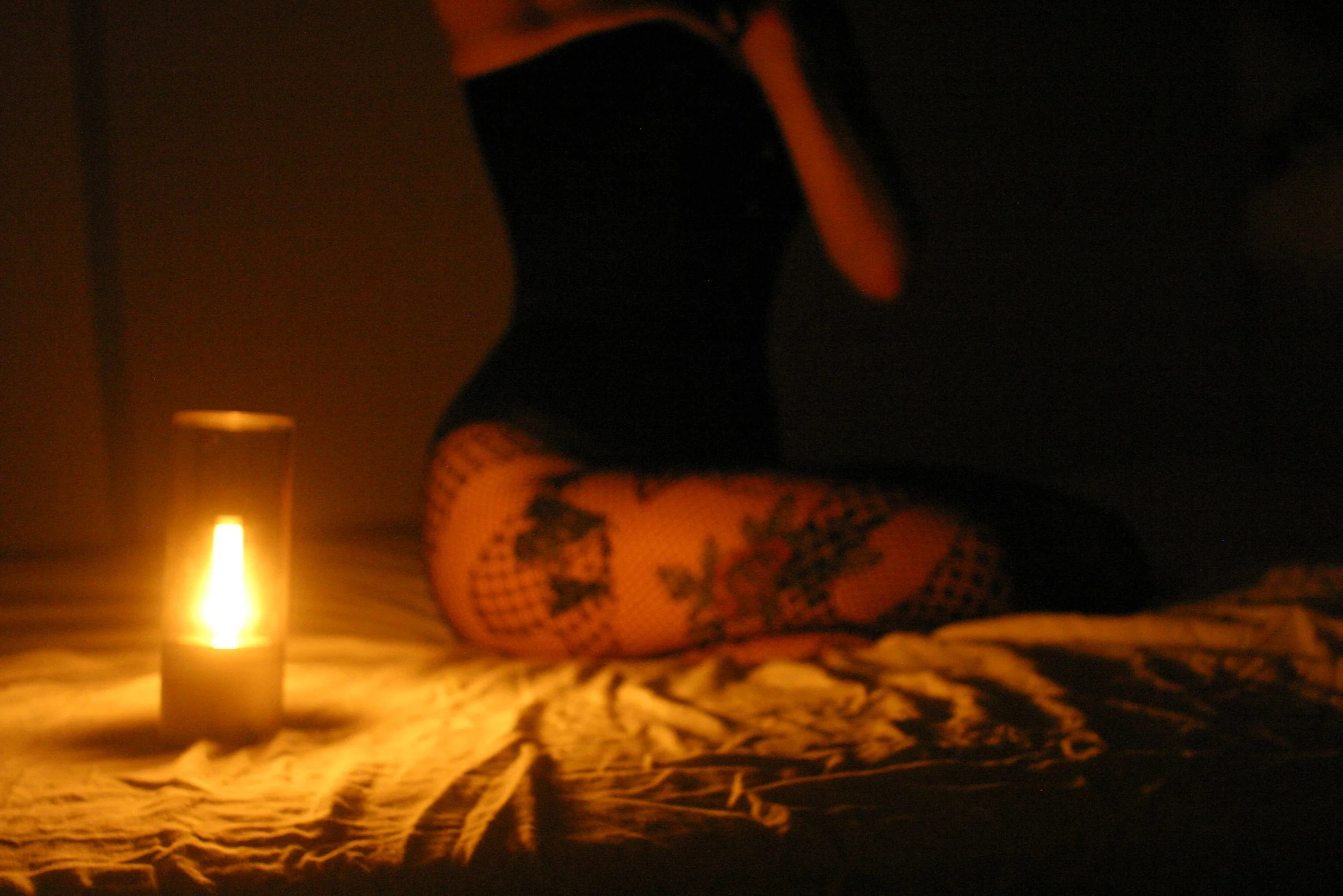 A high-end kinky companion sits on the bed in a black satin corset. Her fishnet tights show off her tattooed thighs. The light is low, and honey-yellow.