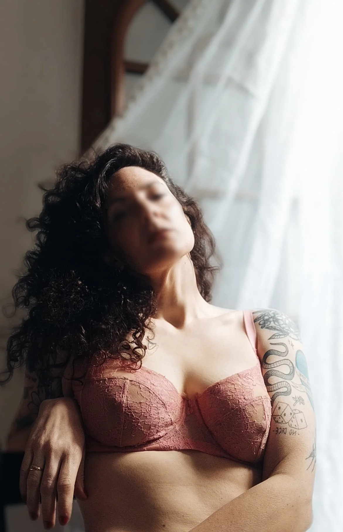 A woman with curly dark hair posing in front of a window with white curtains, wearing a pink lace bra and showing tattoos on her arm.