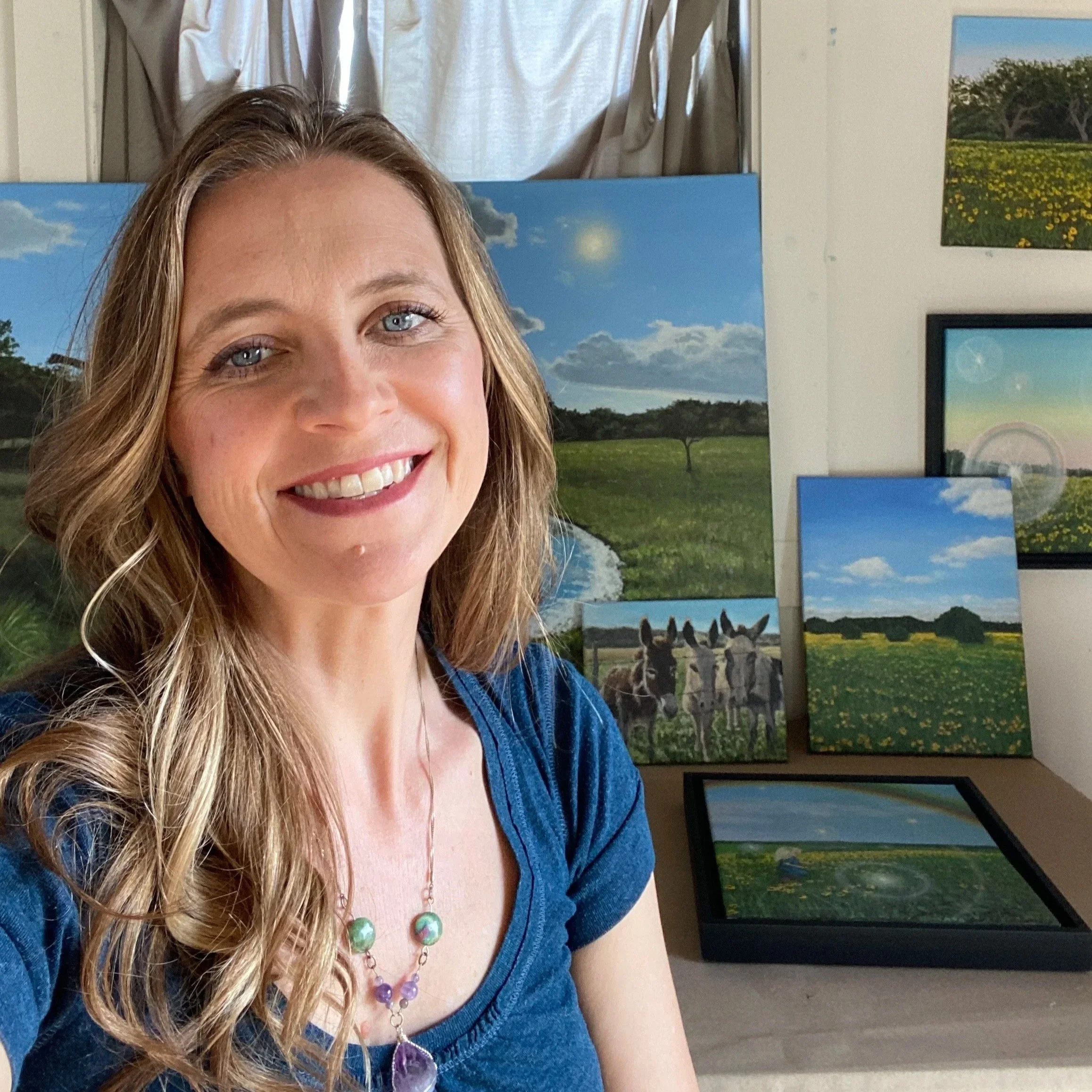 A woman with wavy blonde hair and blue eyes smiling, wearing a blue top and a necklace with large purple and green beads, standing in front of a wall decorated with landscape photographs.