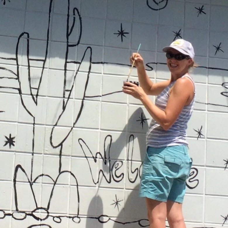 A person painting a mural on a white brick wall. The mural includes a large cactus, some stars, and partly visible text that says "Well".
