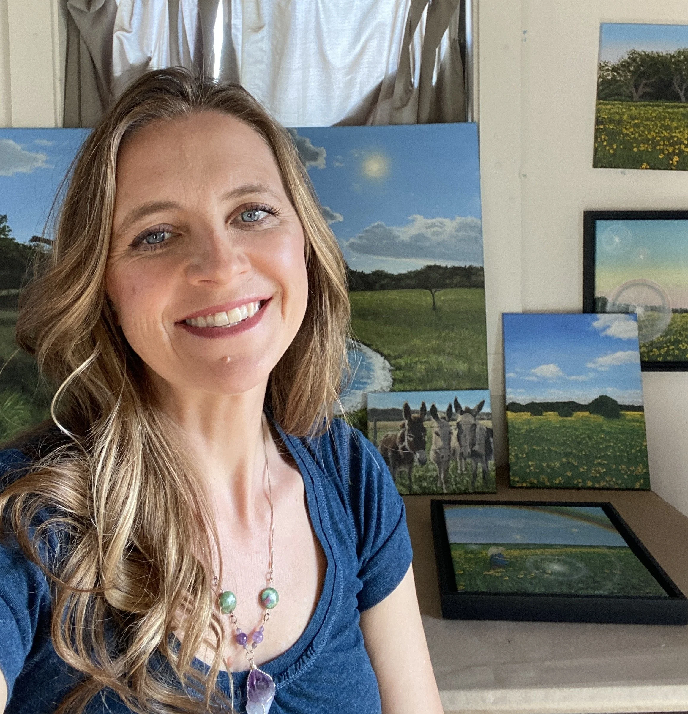 Partial view of a smiling woman with wavy blonde hair, wearing a blue top and colorful jewelry, in front of a wall displaying landscape photographs.
