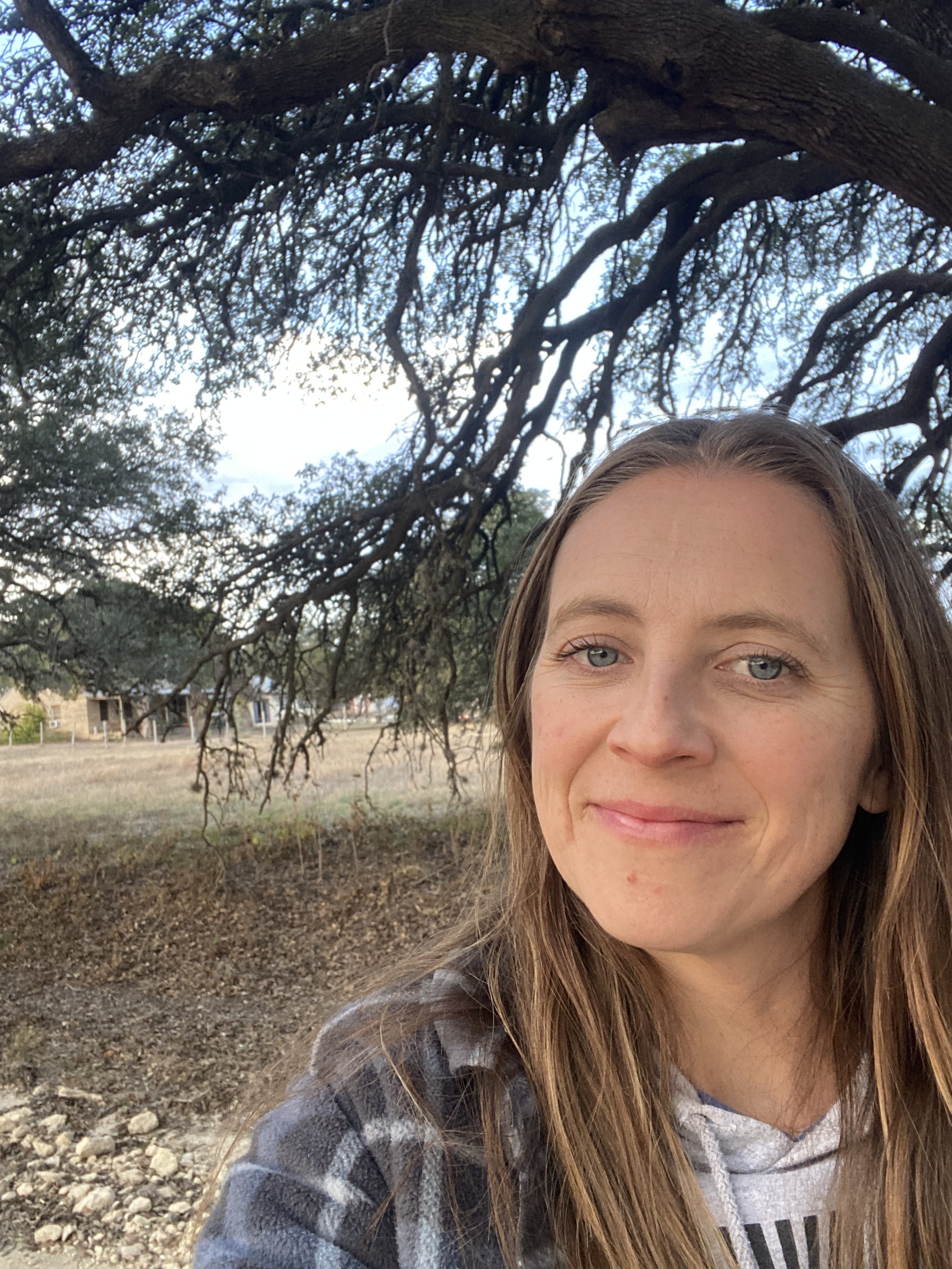 A woman with long light brown hair and blue eyes taking a selfie outdoors, standing next to a large tree with sprawling branches and leaves, with a background of a park or open field.