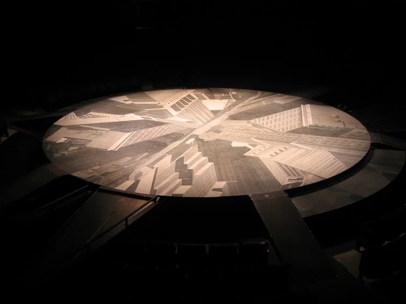 Circular art installation of cityscape buildings and streets, viewed from above in dark lighting.