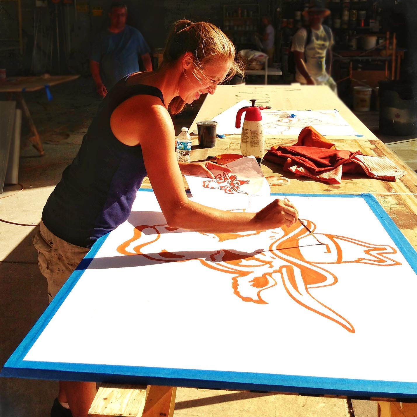 A woman creating a large orange and white artwork of a UT longhorn on a table, outdoors in a workshop setting, with art supplies and other people in the background.