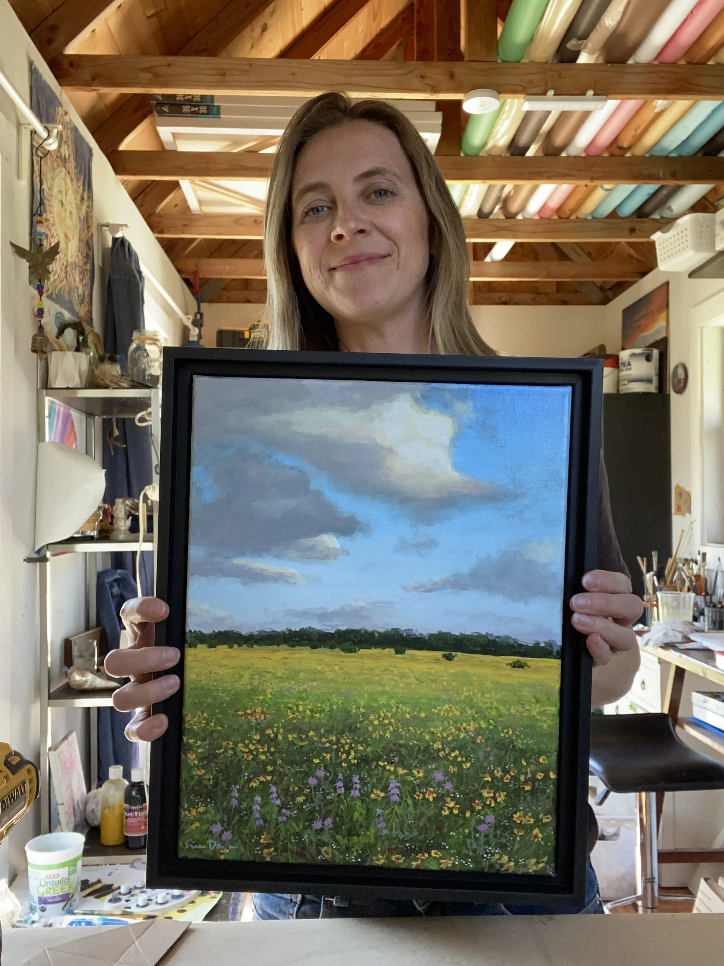 Woman holding a framed landscape painting of a cloudy sky and a colorful field, in a room with art supplies and a work desk.