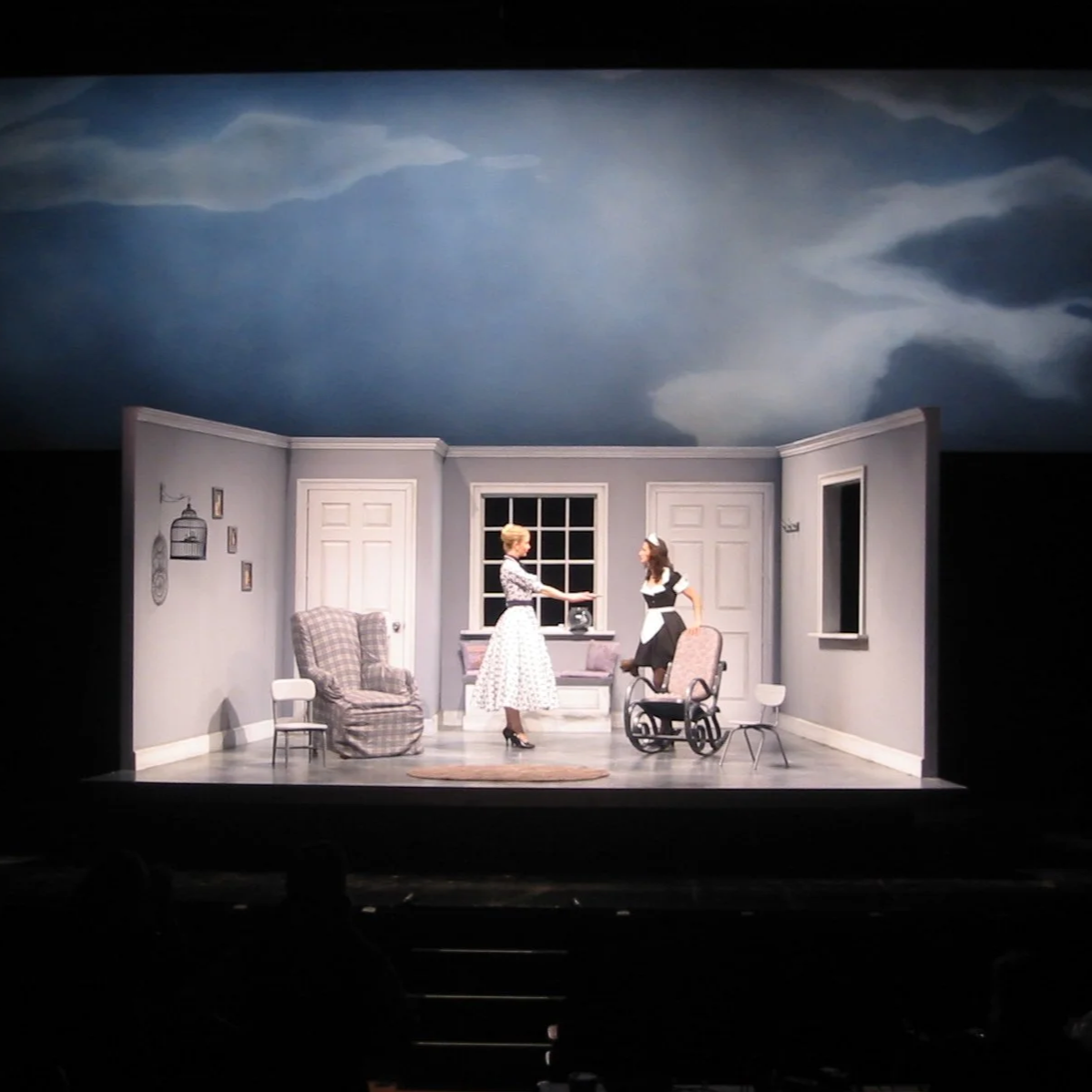 Stage scene of a play set in a vintage-style room with gray walls and a large window. Two women, one in a wheelchair, are interacting, one extending her hand to the other. The room has a plaid armchair, a small chair, and wall decorations. The ceiling has a painted sky with clouds.