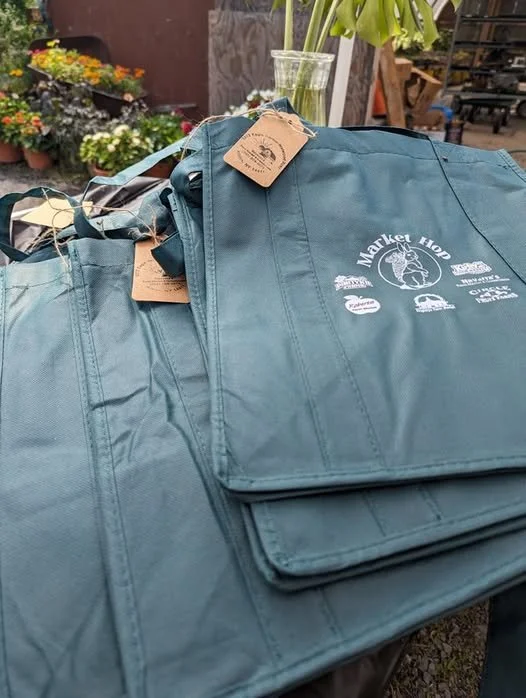 Blue reusable market bags with a logo that says 'Market Hoor' and various logos and text underneath, placed outdoors near plants and flowers.