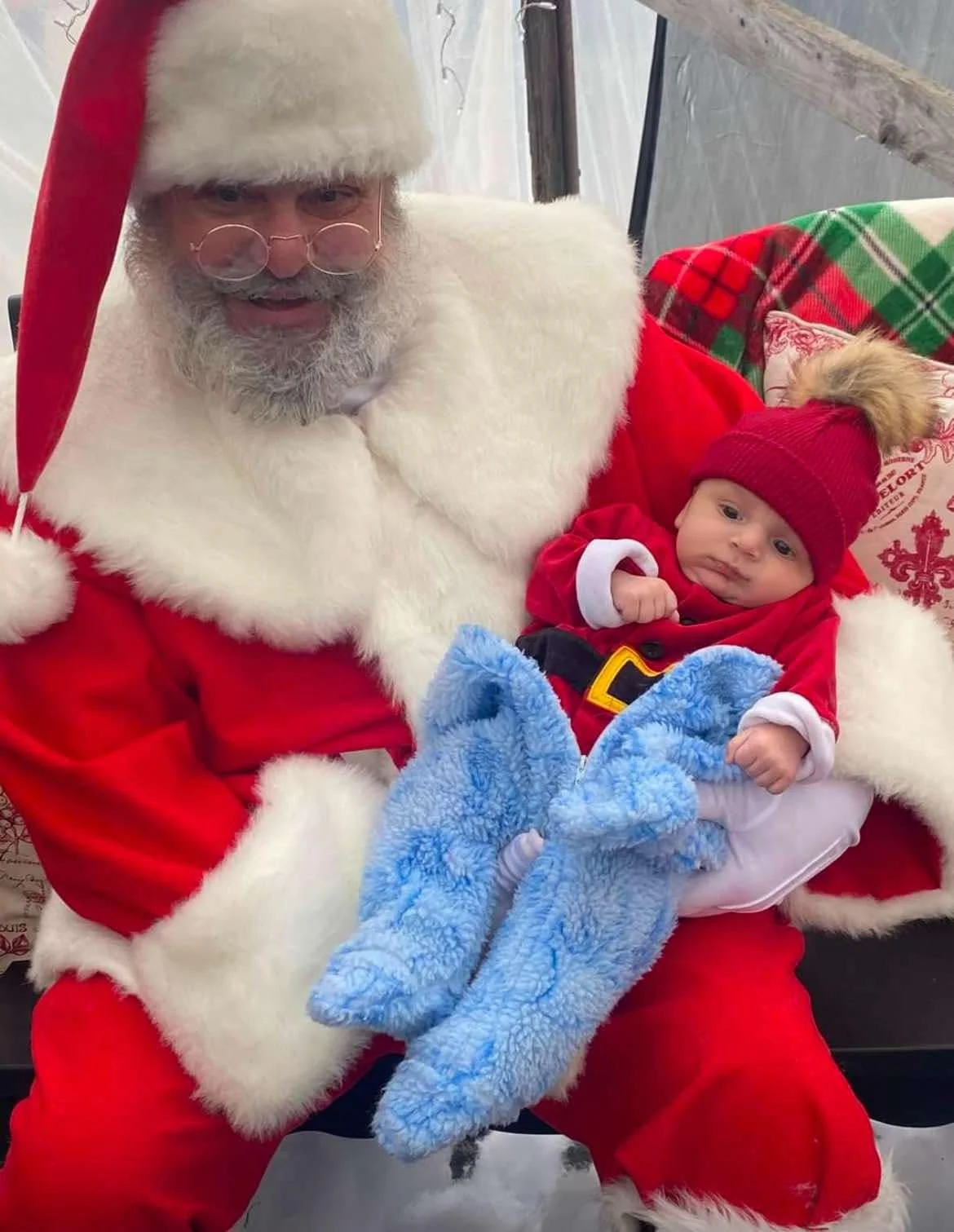 A person dressed as Santa Claus holding a baby in a red and white outfit with a red knit hat. They are sitting in front of a festive holiday background.