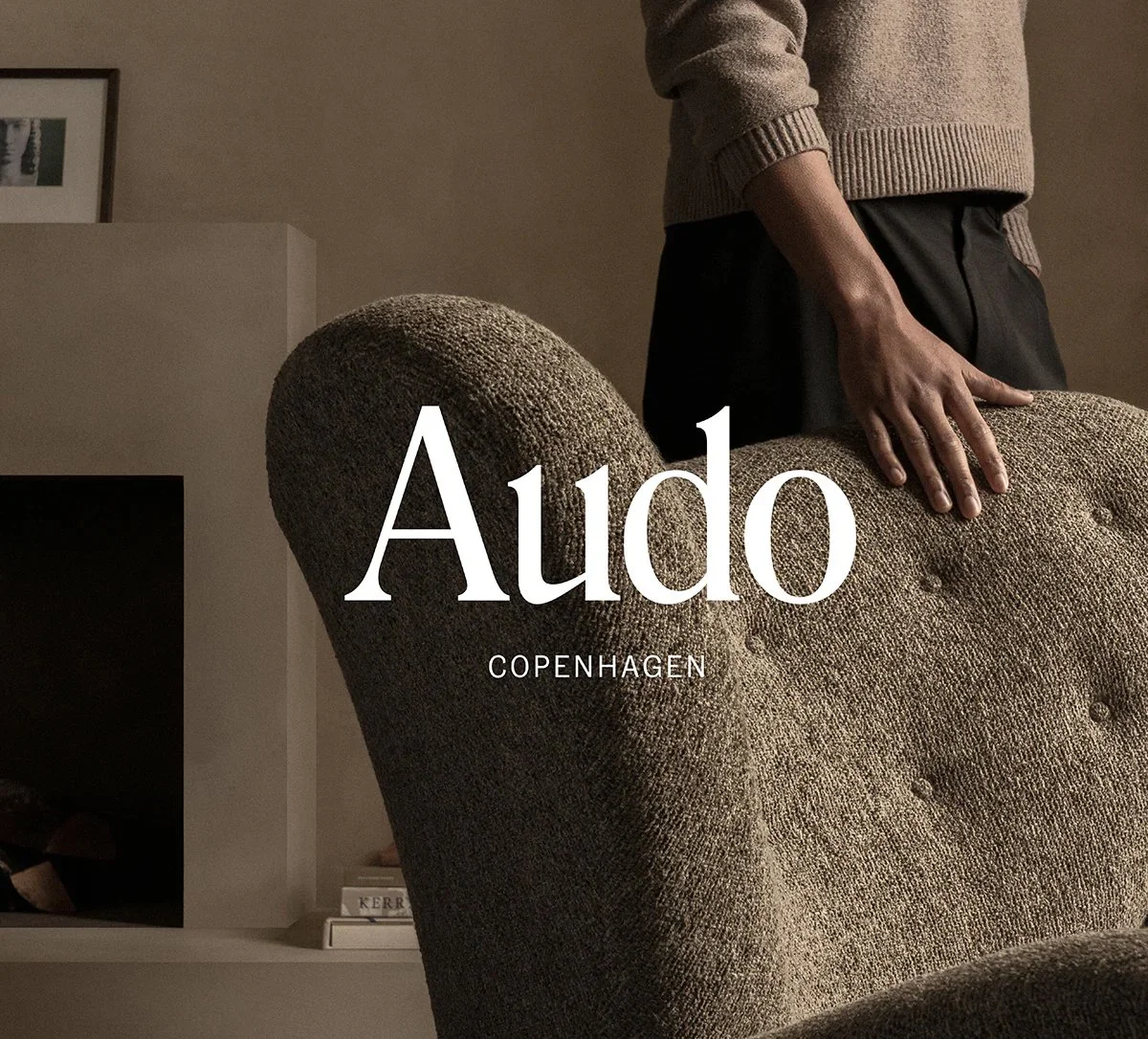 Close-up of a person standing next to a textured brown armchair in a warmly lit room, with the word 'Audo' and 'Copenhagen' overlayed.