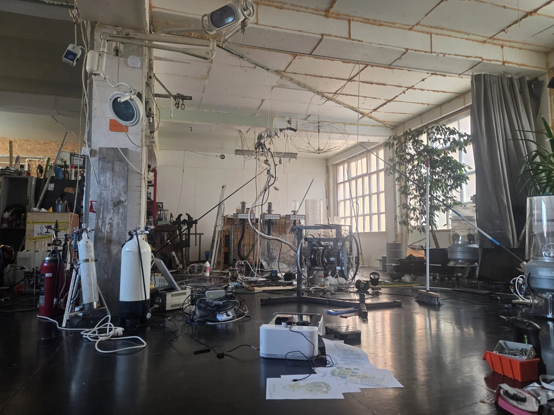 Un atelier de laboratoire ou d'expérimentation avec divers appareils, câbles, bouteilles de gaz, et matériel scientifique, dans un espace iluminé par une grande fenêtre avec des rideaux gris et des plantes en arrière-plan.