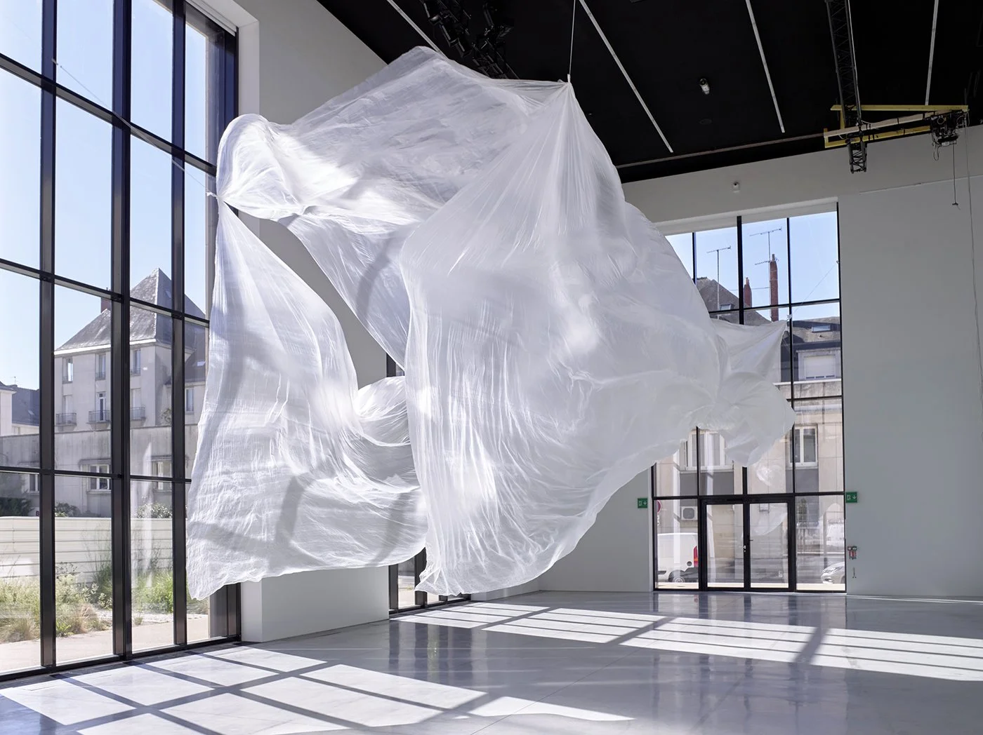 Une sculpture en tissu blanc suspendue dans un espace d'exposition lumineux avec de grandes fenêtres.