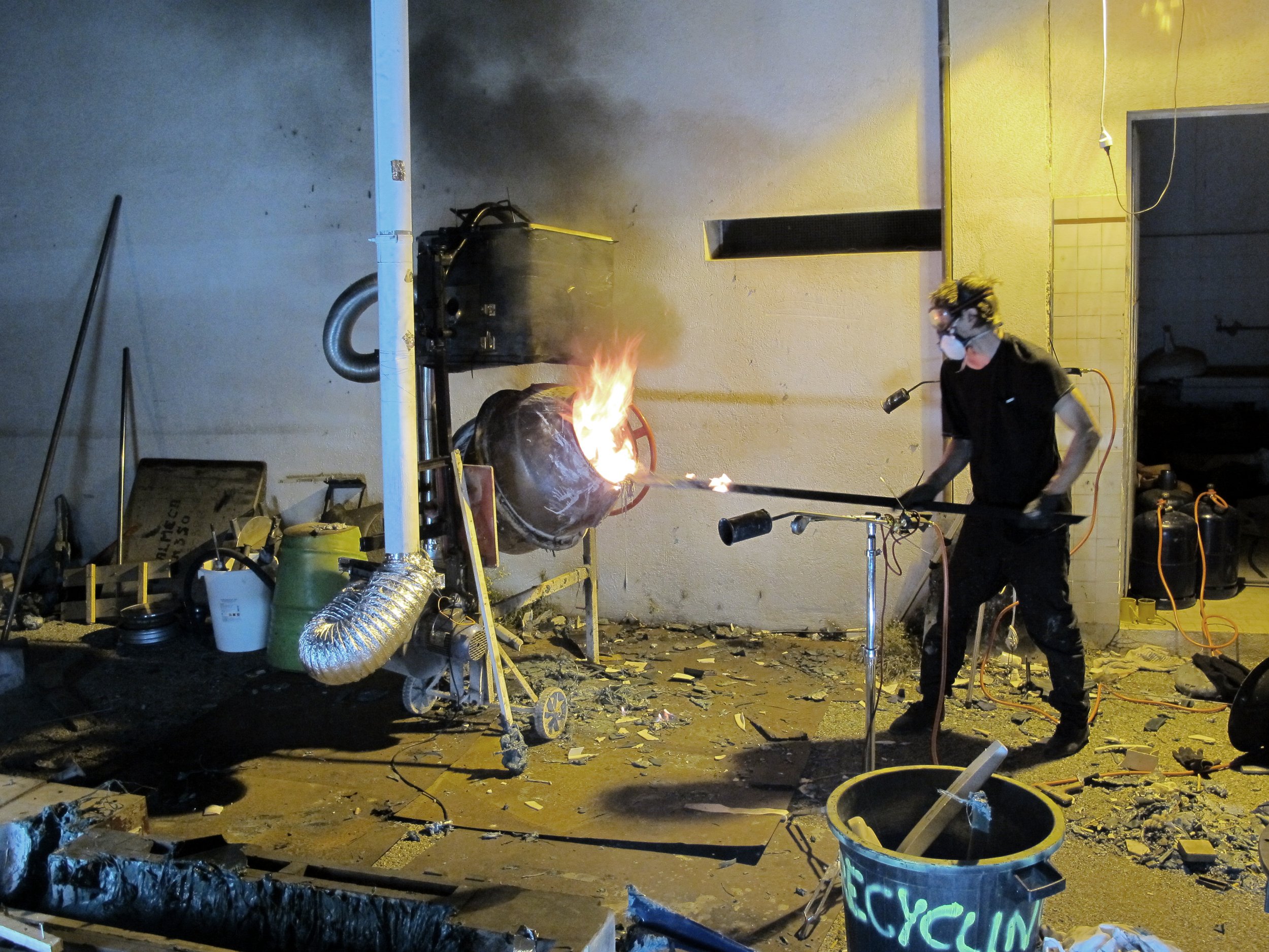 Un homme portant un masque de protection, des lunettes de sécurité et des gants, utilise une torche pour brûler ou couper un objet en feu dans un environnement ressemblant à un atelier ou un chantier, avec des outils, des tuyaux et des déchets autour.