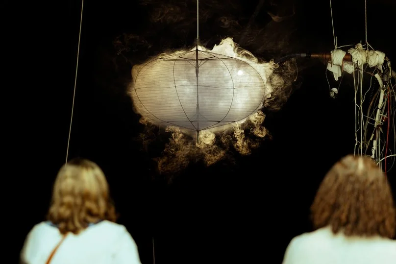 Deux personnes regardant une installation artistique suspendue, composée d'une boule en papier ou en tissu avec de la fumée ou de la vapeur qui en sort, dans un espace sombre.