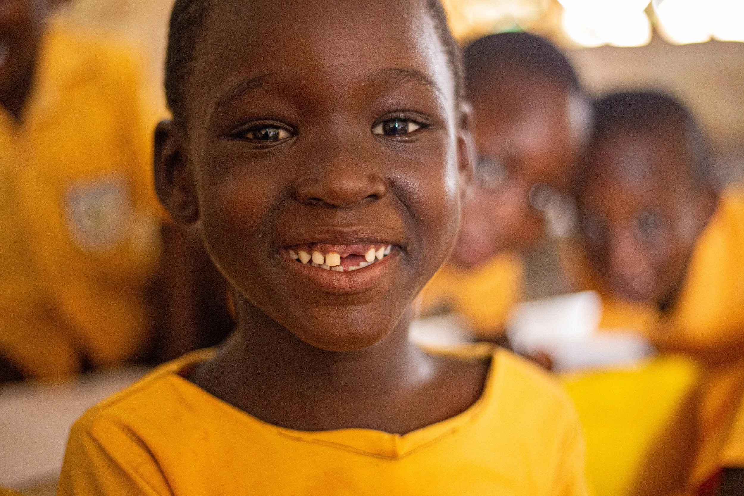 Close-up of a young girl smiling, wearing a yellow shirt, with other children blurred in the background.