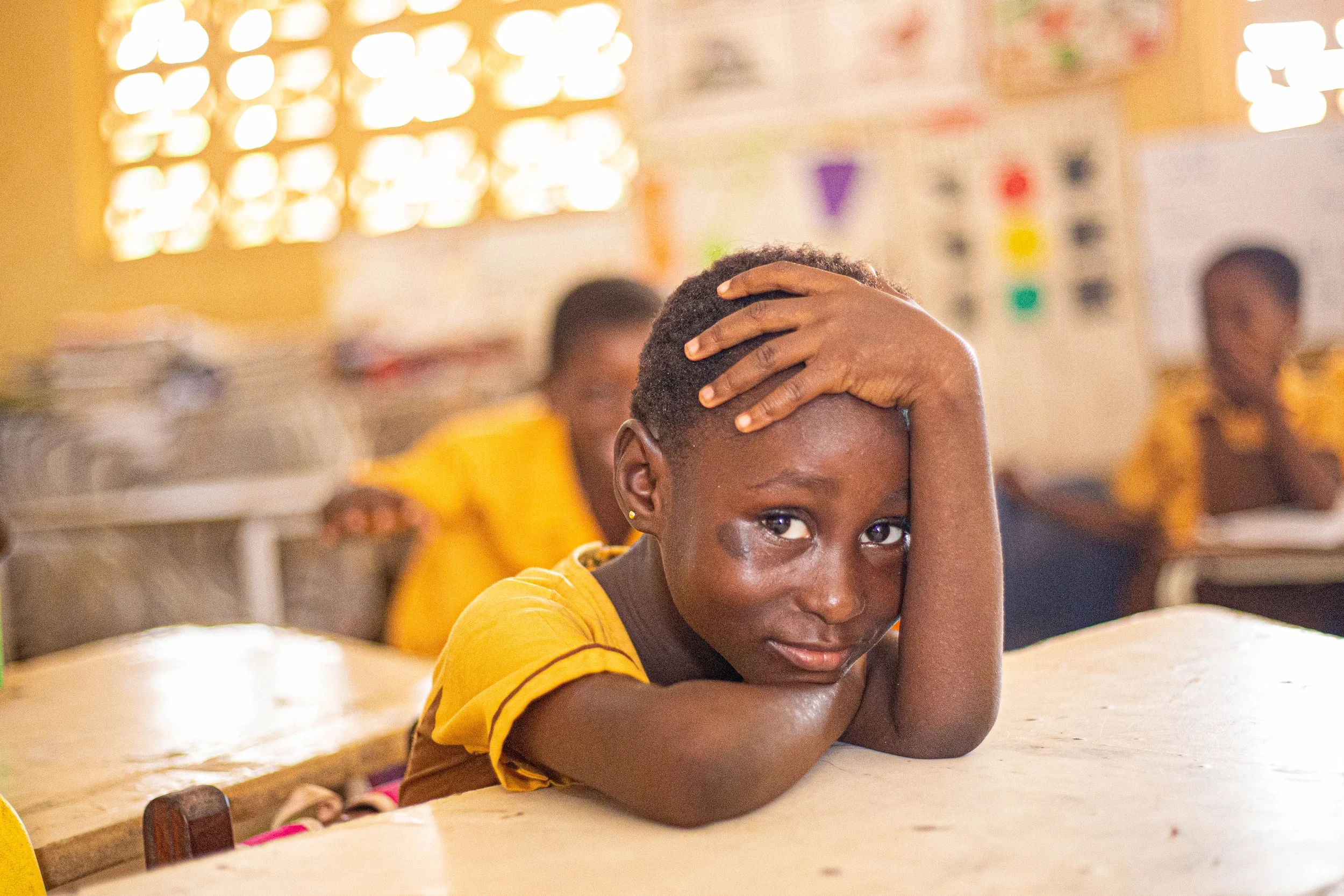 A young girl in a classroom rests her head on her arms on a desk, looking directly at the camera with her hand on her head. The classroom has blurred students and colorful decorations on the wall in the background.
