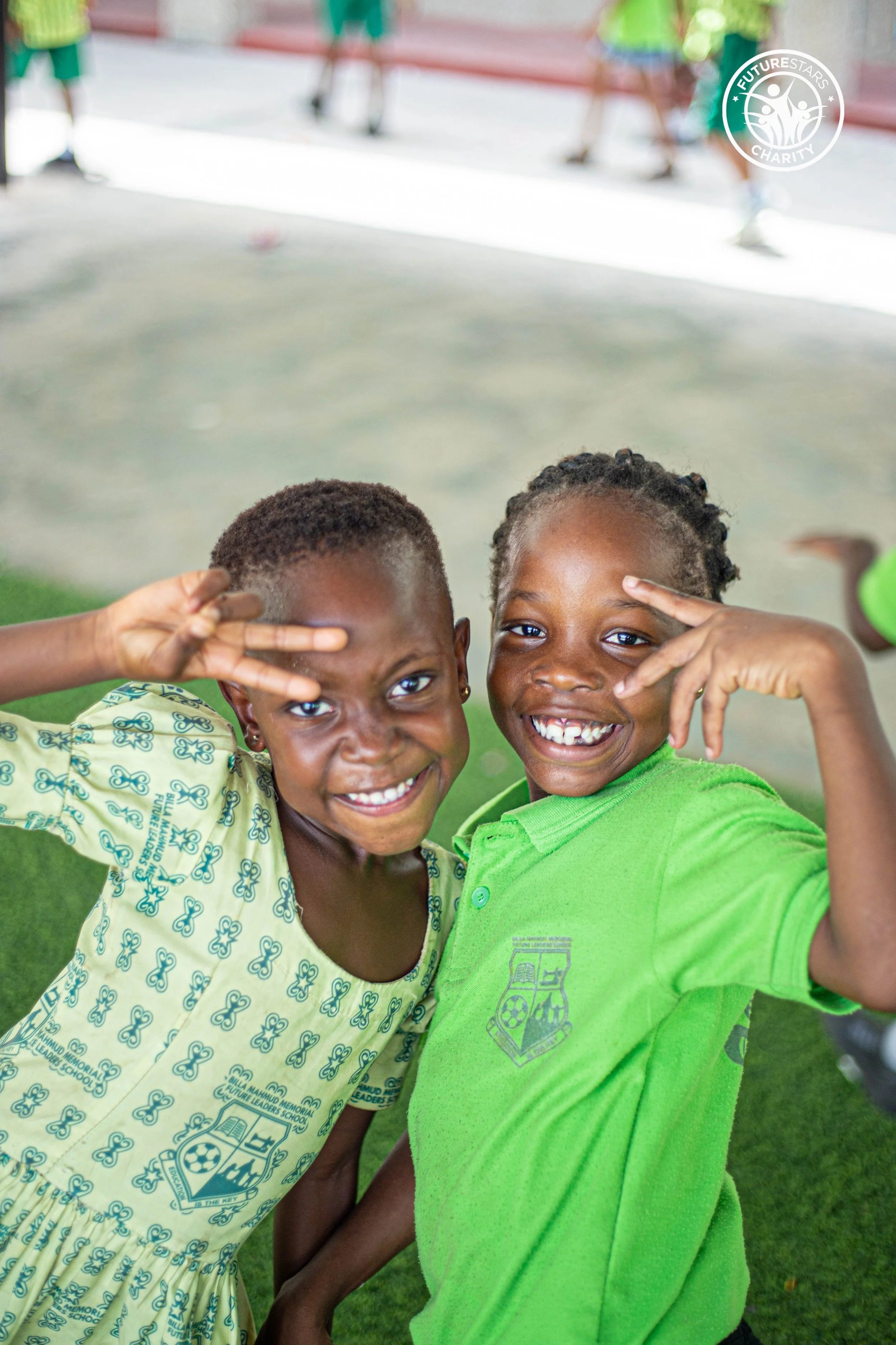 Two smiling children making peace signs with their hands in front of their faces, standing outdoors on a grassy area, with playground equipment and a fence in the background.