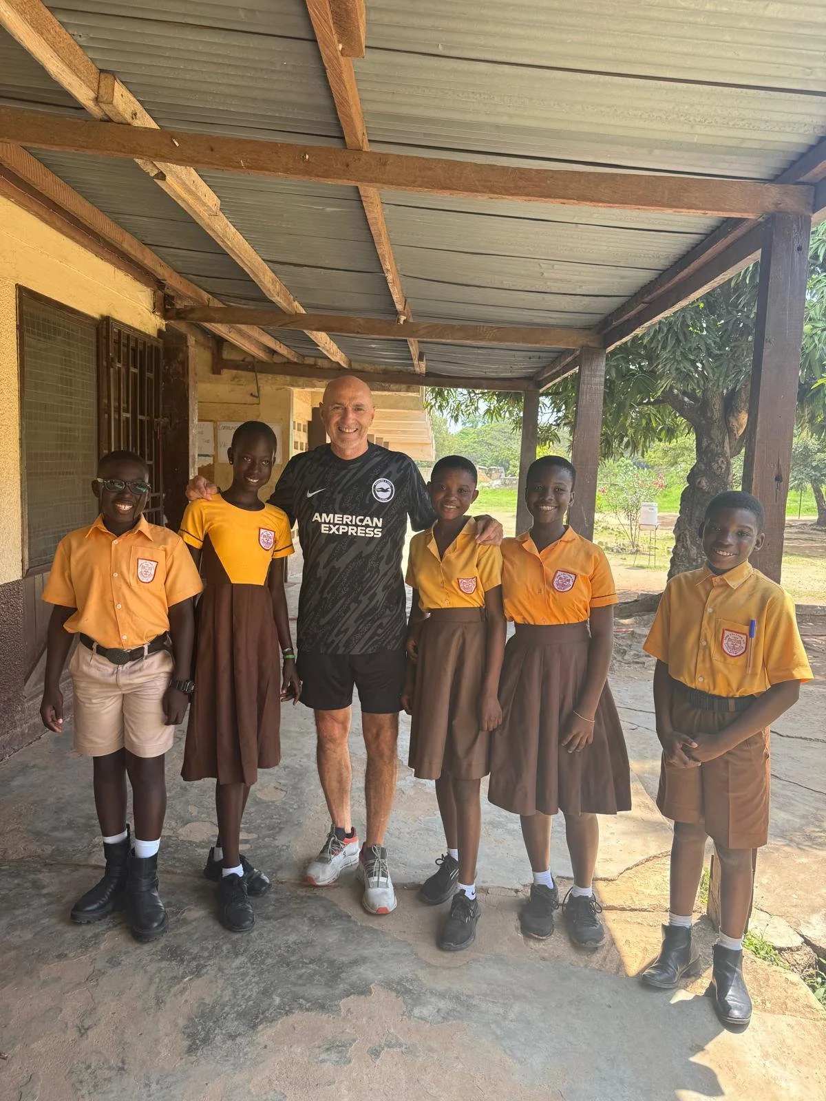 A group of six children and one adult standing outside under a metal roof, smiling at the camera. The children are wearing school uniforms with orange shirts and brown skirts or shorts, and the adult is wearing a black sports jersey with the words 'American Express' on it.