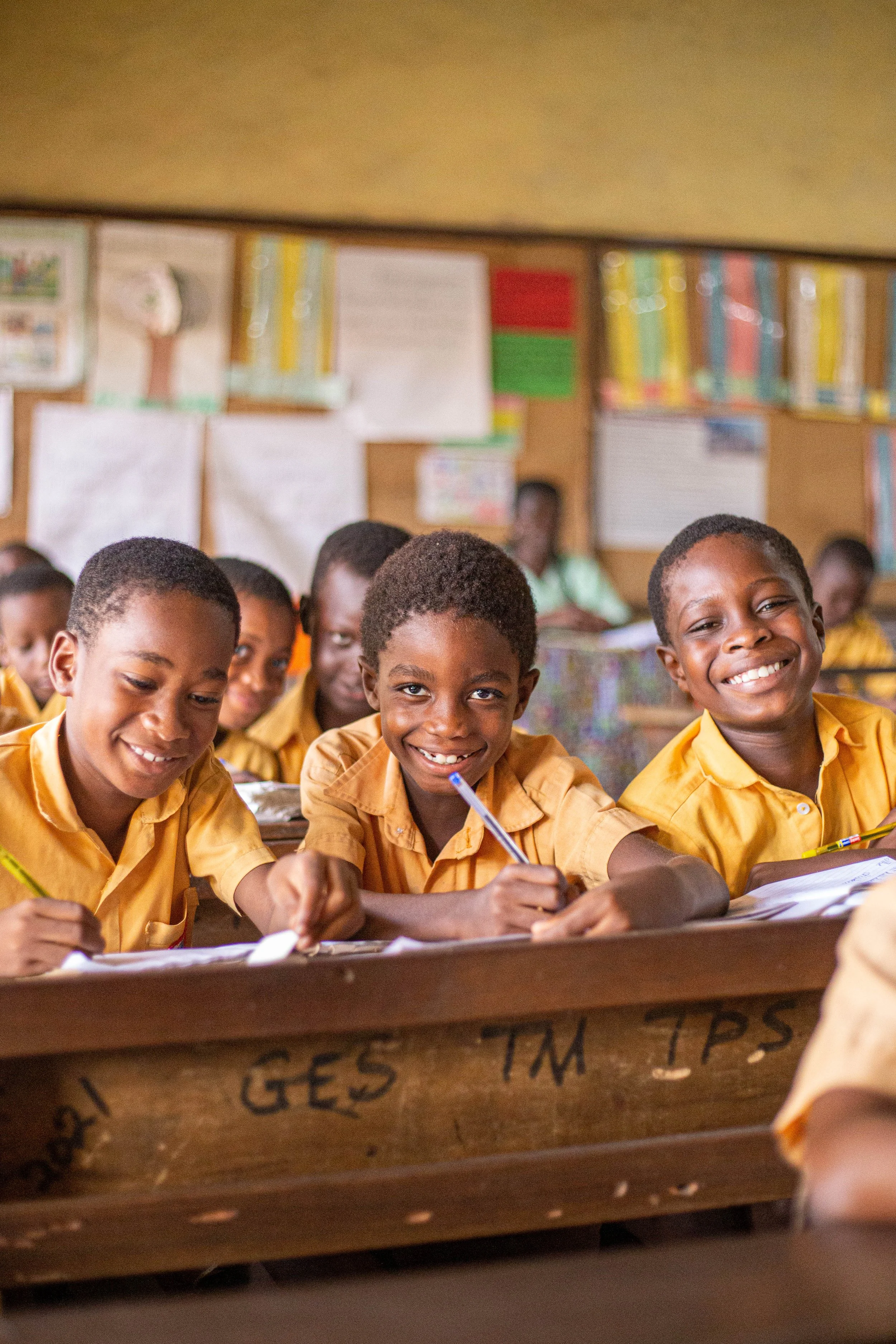 A classroom with young children sitting at desks, smiling, writing, and looking happy. The classroom background includes colorful posters and educational materials on the walls.