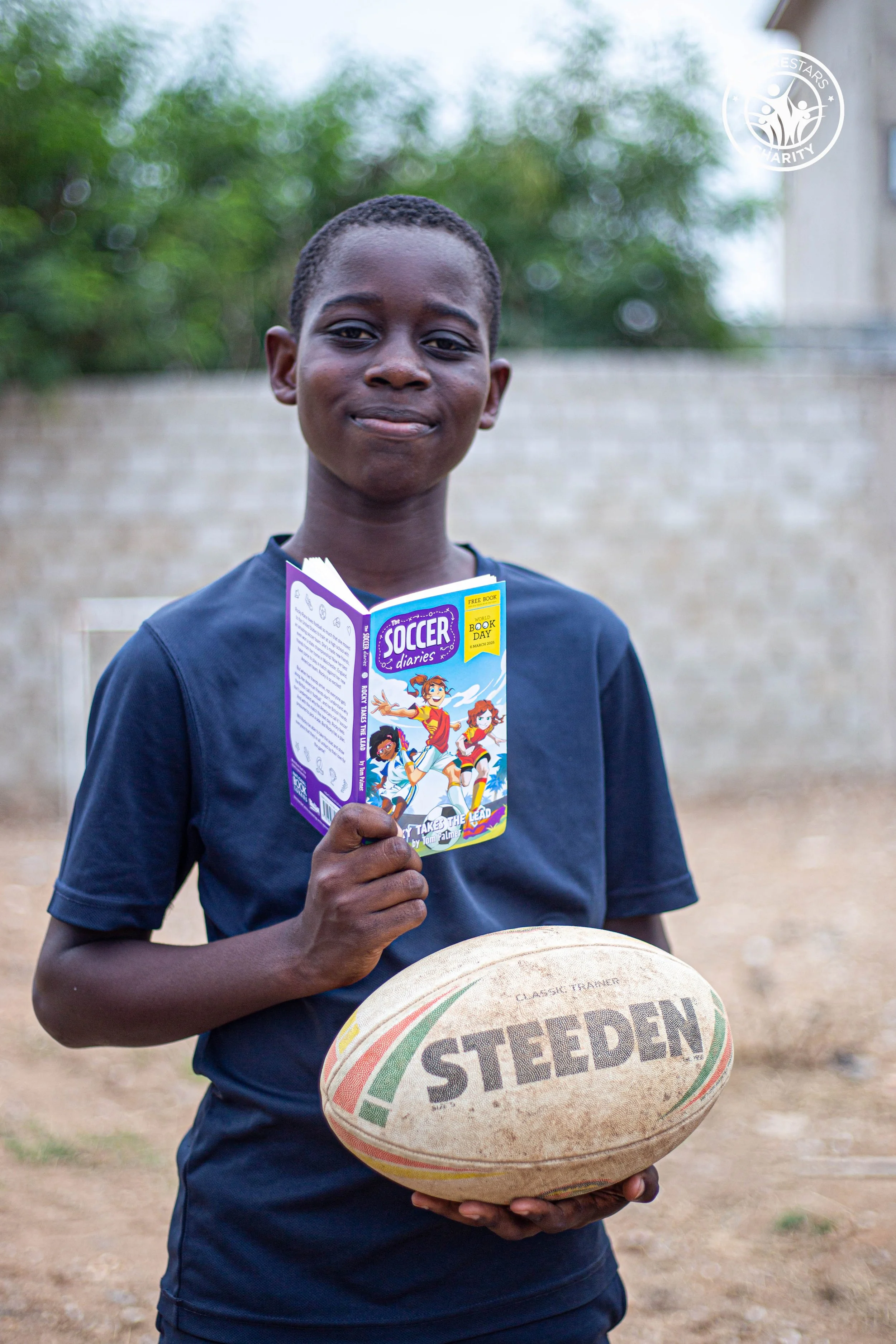 A young boy holding a rugby ball with the word 'STEDEN' on it, a rugby ball, in an outdoor setting. He is also holding a book titled 'Soccer Diaries' with cartoon characters on the cover, and a basketball is partially visible in the foreground. The background shows a beige brick wall and trees.