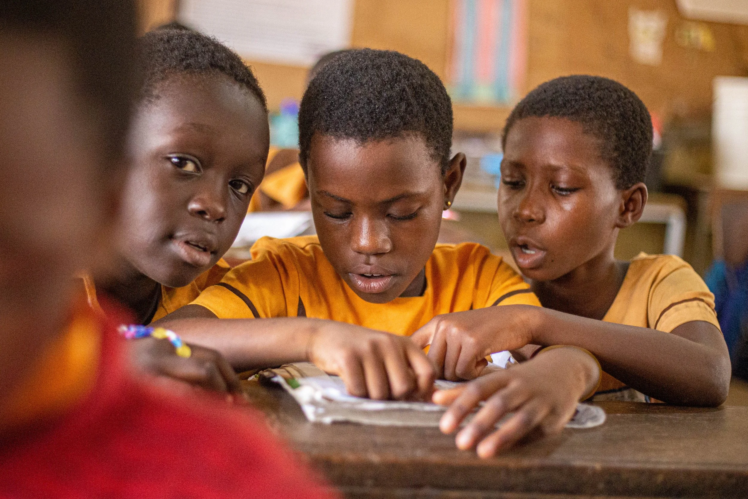 Three children, two boys and one girl, focusing on an activity at a table in a classroom, with classroom decorations in the background.