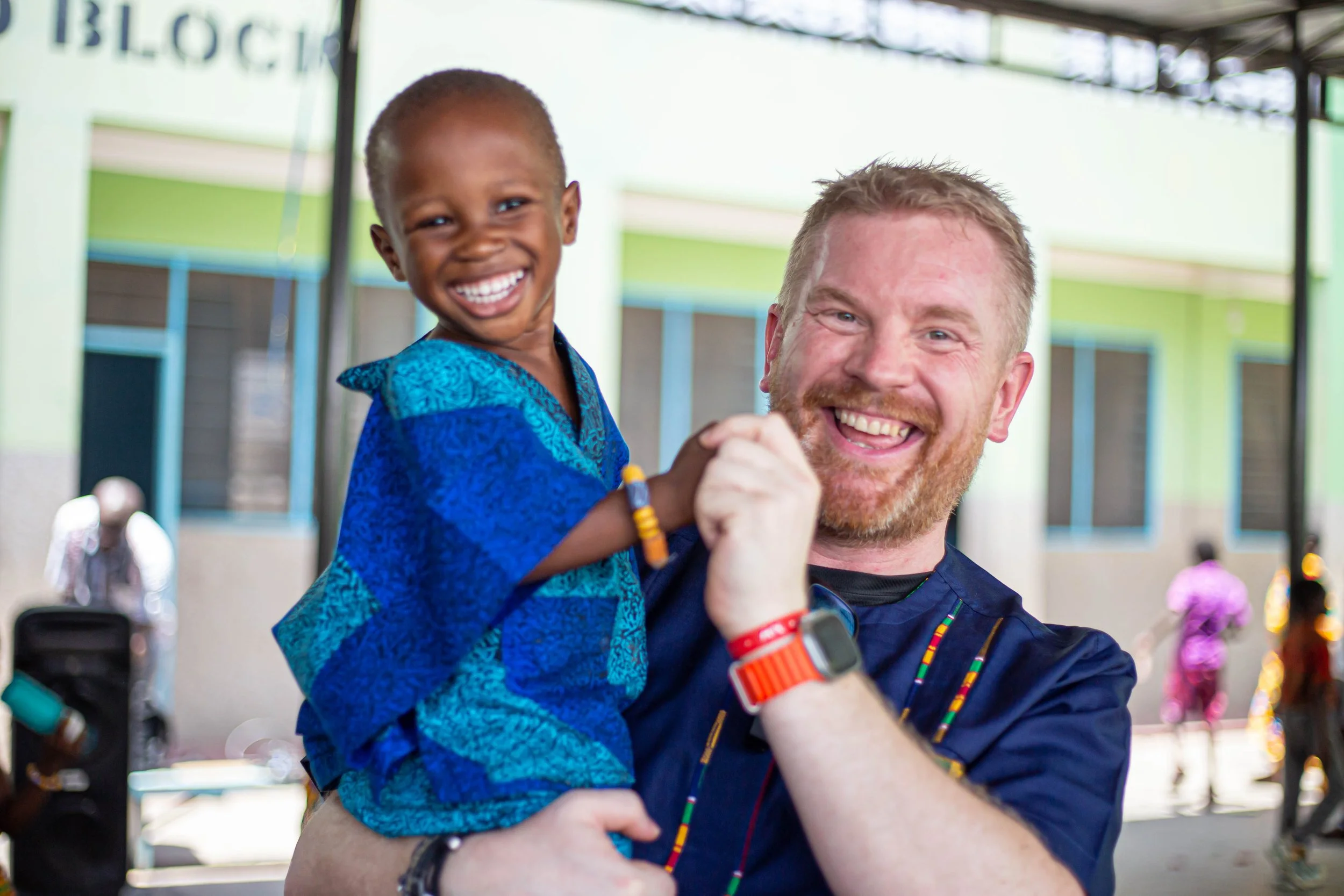 A young boy wearing a blue shirt smiling and being held by a man with a beard, smiling, at an outdoor event.